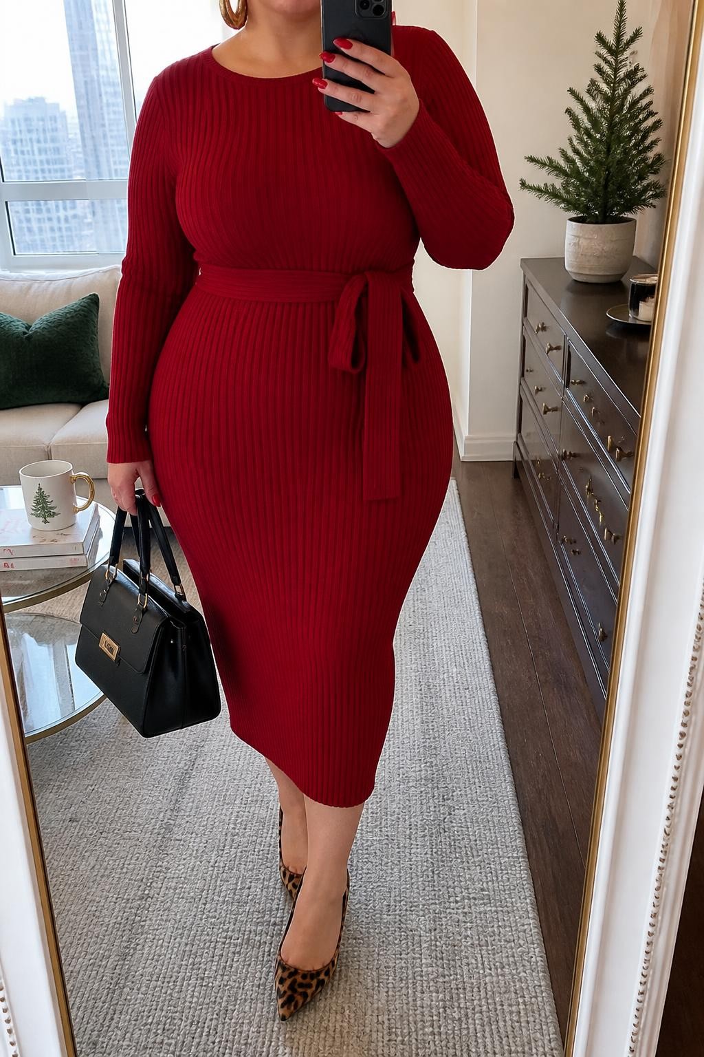 Mirror selfie of a plus size woman wearing a ruby red ribbed sweater midi dress with long sleeves and a matching waist tie, leopard print pointed pumps, a black structured handbag, and chunky gold hoop earrings, clearly showing the exact clothing items, red ribbed texture, waist tie, print, colors, and accessories listed, holding a phone in front of a mirror, cropped at the neck so the face does not show, relaxed confident selfie pose, in a stylish city apartment with natural window light, a coffee mug on a side table and a small plant on a dresser, realistic festive daytime styling, latest iPhone photo quality.