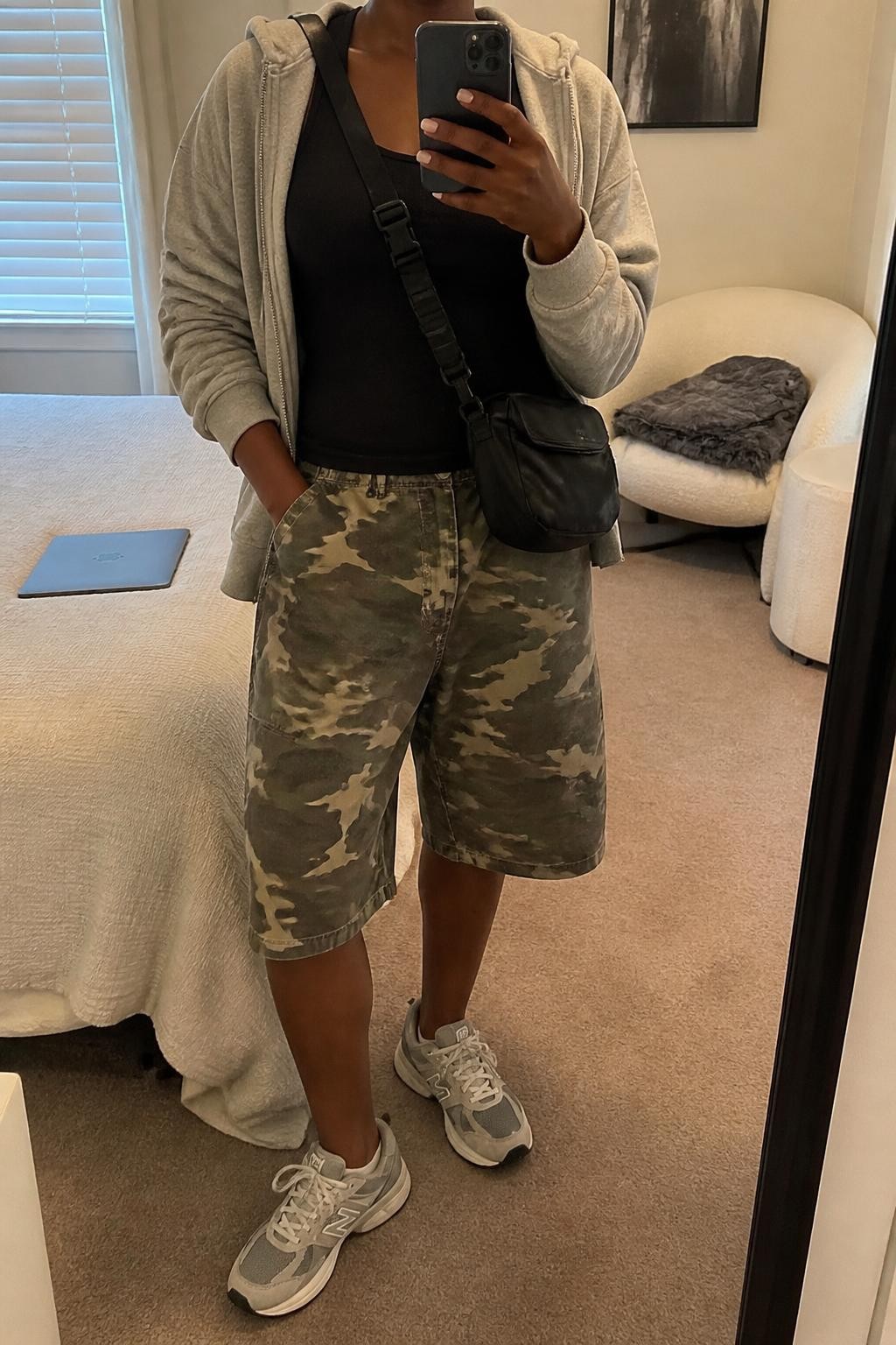 mirror selfie of a Black woman wearing oversized woodland camouflage denim jorts, a heather gray zip hoodie worn open over a fitted black cotton tank, gray and white athletic sneakers and a black nylon crossbody bag, wearing exactly the listed clothing and accessories with no additional garments, holding a phone in front of a mirror, cropped at NECK so the face does not show, relaxed sporty selfie pose, in a cozy bedroom mirror setup, soft indoor lighting, laptop on the bed and a folded blanket on a chair, realistic everyday latest iPhone photo quality.