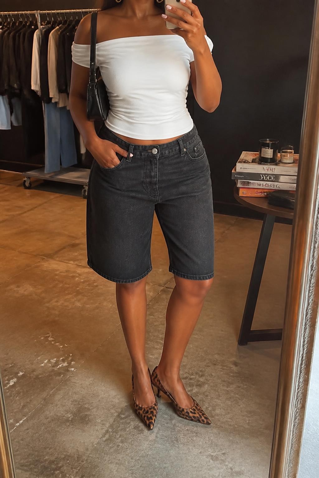 mirror selfie of a Black woman wearing faded black denim jorts with a relaxed knee-length cut, a fitted white off-shoulder stretch cotton top, leopard print pointed kitten heels in warm brown and black tones, a black mini shoulder bag and medium gold hoop earrings, wearing exactly the listed outfit without additional garments, holding a phone in front of a mirror, cropped at NECK so the face does not show, relaxed confident selfie pose, in a fashion studio mirror corner, warm moody lighting, clothing rack with garments behind and a stack of fashion magazines on a side table, natural unstaged styling, latest iPhone photo quality.