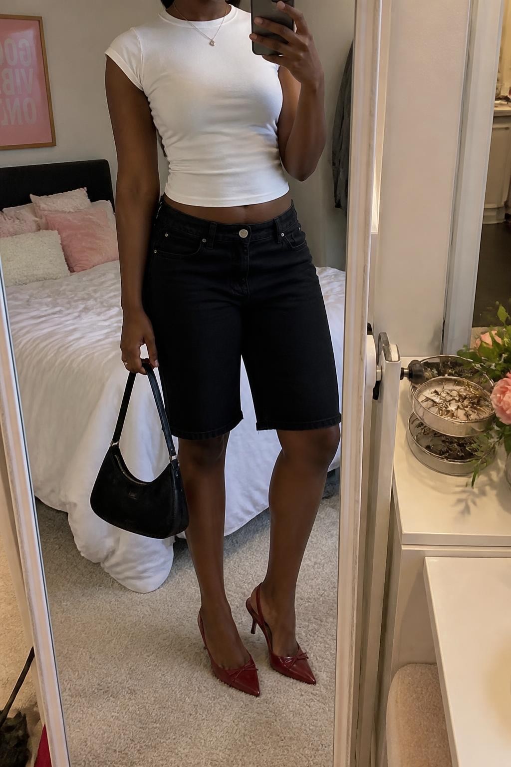mirror selfie of a Black woman wearing black Bermuda denim jorts with a clean straight fit, a fitted white cotton baby tee, cherry red pointed kitten heels, a small black leather handbag and a thin silver pendant necklace, wearing exactly the listed outfit with no extra clothing pieces, holding a phone in front of a mirror, cropped at NECK so the face does not show, relaxed casual selfie pose, in a feminine-themed bedroom, soft indoor lighting, jewelry tray on the vanity and soft pillows on an unmade bed, believable everyday styling, latest iPhone photo quality.