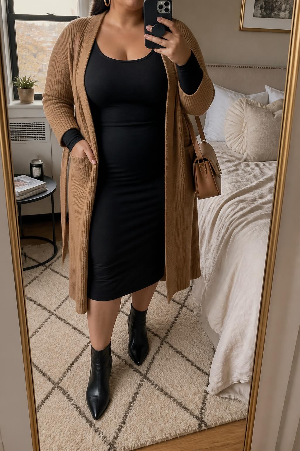 Mirror selfie of a plus size woman wearing a fitted black long-sleeve jersey midi dress with a scoop neckline, a camel knit coatigan worn open with its matching belt tied loosely behind the waist, black pointed ankle boots, a camel leather shoulder bag, and chunky gold hoop earrings, clearly showing the exact clothing items, black jersey dress, camel knit layer, belt placement, footwear, and accessories listed, holding a phone in front of a mirror, cropped at the neck so her face does not appear, relaxed sophisticated selfie stance, in a cozy bedroom mirror setup with natural window light, soft pillows on an unmade bed and a stack of fashion magazines on a side table, realistic cold-weather Thanksgiving styling, latest iPhone photo quality.