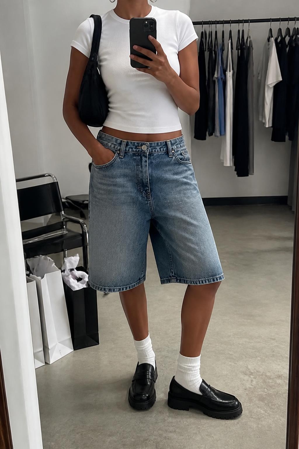 mirror selfie of a Black woman wearing baggy medium-wash blue denim jorts with a loose knee-length cut, a fitted white cotton baby tee, chunky black leather loafers with a thick sole, white ribbed crew socks, a black nylon shoulder bag and medium silver hoop earrings, wearing exactly the listed clothing and accessories with nothing additional, holding a phone in front of a mirror, cropped at NECK so the face does not show, relaxed cool-girl selfie pose, in a fashion studio mirror corner, soft indoor lighting, clothing rack with garments behind and shopping bags beside a chair, realistic casual proportions, latest iPhone photo quality.
