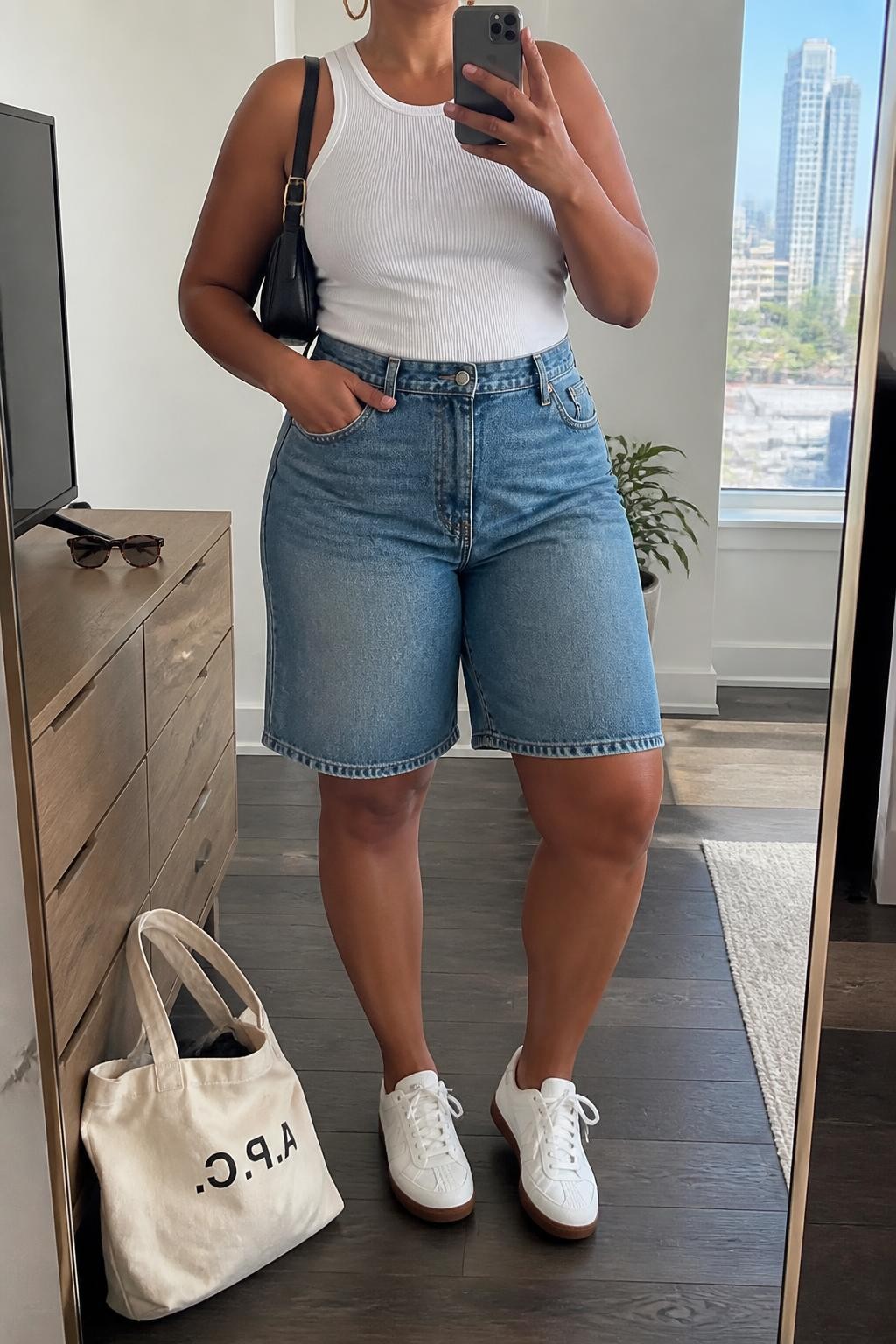 mirror selfie of a plus size Black woman wearing high-waisted medium-wash blue denim jorts with a relaxed knee-length fit, a fitted white ribbed cotton tank top, white retro sneakers, a black leather shoulder bag and medium gold hoop earrings, wearing exactly the listed clothing and accessories with no extra garments, holding a phone in front of a mirror, cropped at NECK so the face does not show, relaxed confident selfie pose, in a stylish city apartment, natural window light, sunglasses placed on a dresser and a tote bag resting on the floor, realistic body proportions and natural denim folds, latest iPhone photo quality.