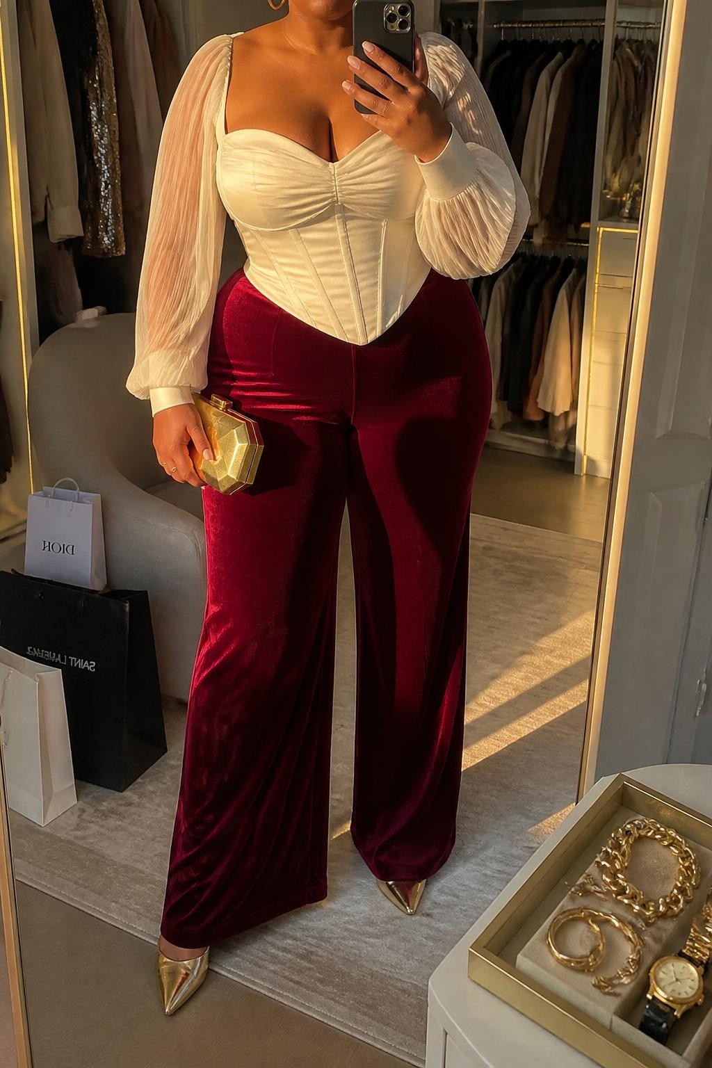 Mirror selfie of a plus size woman wearing burgundy velvet wide-leg pants, an ivory satin corset-style blouse with long sheer sleeves and a structured opaque bodice, gold pointed heels, a gold hard-case clutch, and delicate gold hoop earrings, clearly showing the exact clothing items, burgundy velvet, ivory satin, sheer sleeves, colors, footwear, and accessories listed, holding a phone in front of a mirror, cropped at the neck so the face does not show, relaxed glamorous selfie pose, in a walk-in closet mirror with evening golden-hour lighting, shopping bags beside a chair and a jewelry tray on the vanity, realistic show-stopping Christmas event appearance, latest iPhone photo quality.