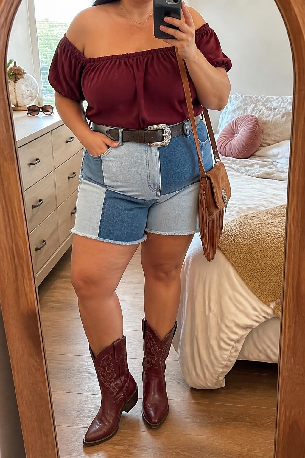 Mirror selfie of a plus size woman wearing high-waisted patchwork denim shorts with mixed light and medium blue panels and a relaxed mid-thigh length, a burgundy off-shoulder blouse with short gathered sleeves, burgundy leather mid-calf cowboy boots, a dark brown Western belt with a silver buckle, and a small tan fringe crossbody bag, clearly showing the exact clothing items, mixed denim panels, burgundy blouse, matching boots, belt, and accessories listed, holding a phone in front of a mirror, cropped at the neck so the face does not appear, relaxed playful selfie stance, in a cozy bedroom mirror setup with natural window light, sunglasses placed on a dresser and soft pillows on an unmade bed, realistic summer vaquera outfit, latest iPhone photo quality.