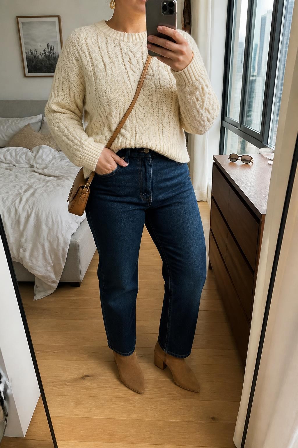 Mirror selfie of a plus size woman wearing dark indigo high-waisted straight-leg jeans, a cream cable-knit sweater with a relaxed fit and slightly cropped hem, tan suede ankle boots, a camel leather crossbody bag, and hammered gold drop earrings, clearly showing the exact clothing items, dark denim, cream cable knit, tan footwear, and accessories listed, holding a phone in front of a mirror, cropped at the neck so her face does not appear, relaxed casual selfie stance, in a stylish city apartment with natural window light, soft pillows on an unmade bed and sunglasses placed on a dresser, realistic family Thanksgiving outfit, latest iPhone photo quality.