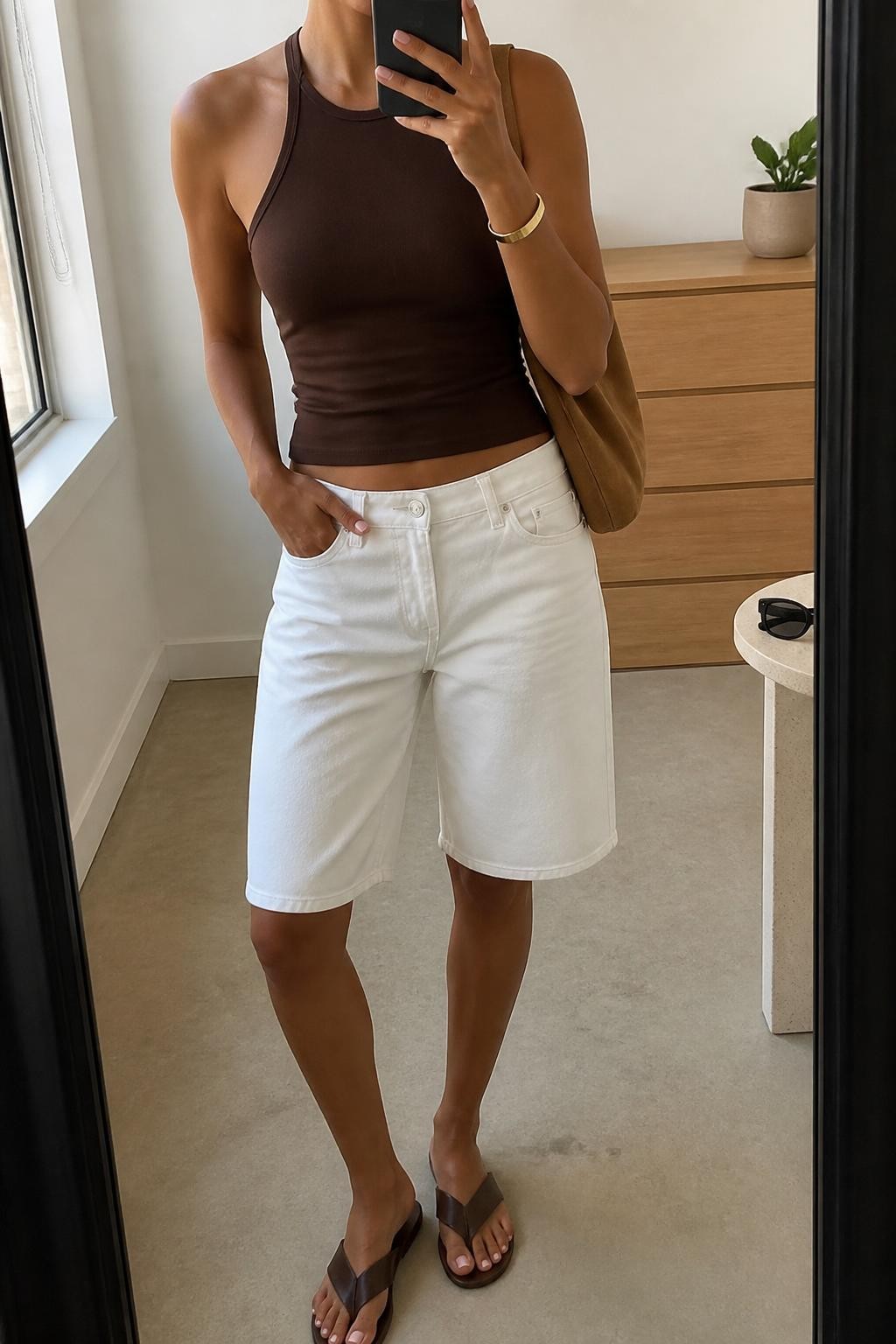 mirror selfie of a woman with medium brown skin wearing white relaxed denim jorts with a straight knee-length fit, a fitted chocolate brown halter top in stretch cotton, brown flat leather sandals, a tan suede shoulder bag and a simple gold cuff bracelet, wearing exactly the listed clothing and accessories with no extras, holding a phone in front of a mirror, cropped at NECK so the face does not show, relaxed casual selfie pose, in a minimal dressing room, natural window light, a small plant on a dresser and sunglasses placed on a side table, natural warm-weather styling, latest iPhone photo quality.