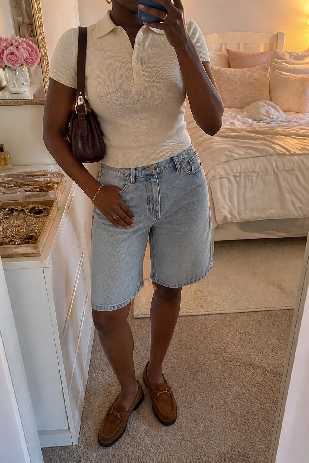mirror selfie of a Black woman wearing light-wash relaxed denim jorts with a knee-length fit, a fitted cream short-sleeve fine-knit polo, tan brown suede loafers, a dark brown leather shoulder bag and medium gold hoop earrings, wearing exactly the listed outfit with no extra clothing pieces, holding a phone in front of a mirror, cropped at NECK so the face does not show, relaxed polished selfie pose, in a feminine-themed bedroom, evening golden hour, soft pillows on an unmade bed and a jewelry tray on the vanity, believable casual styling, latest iPhone photo quality.