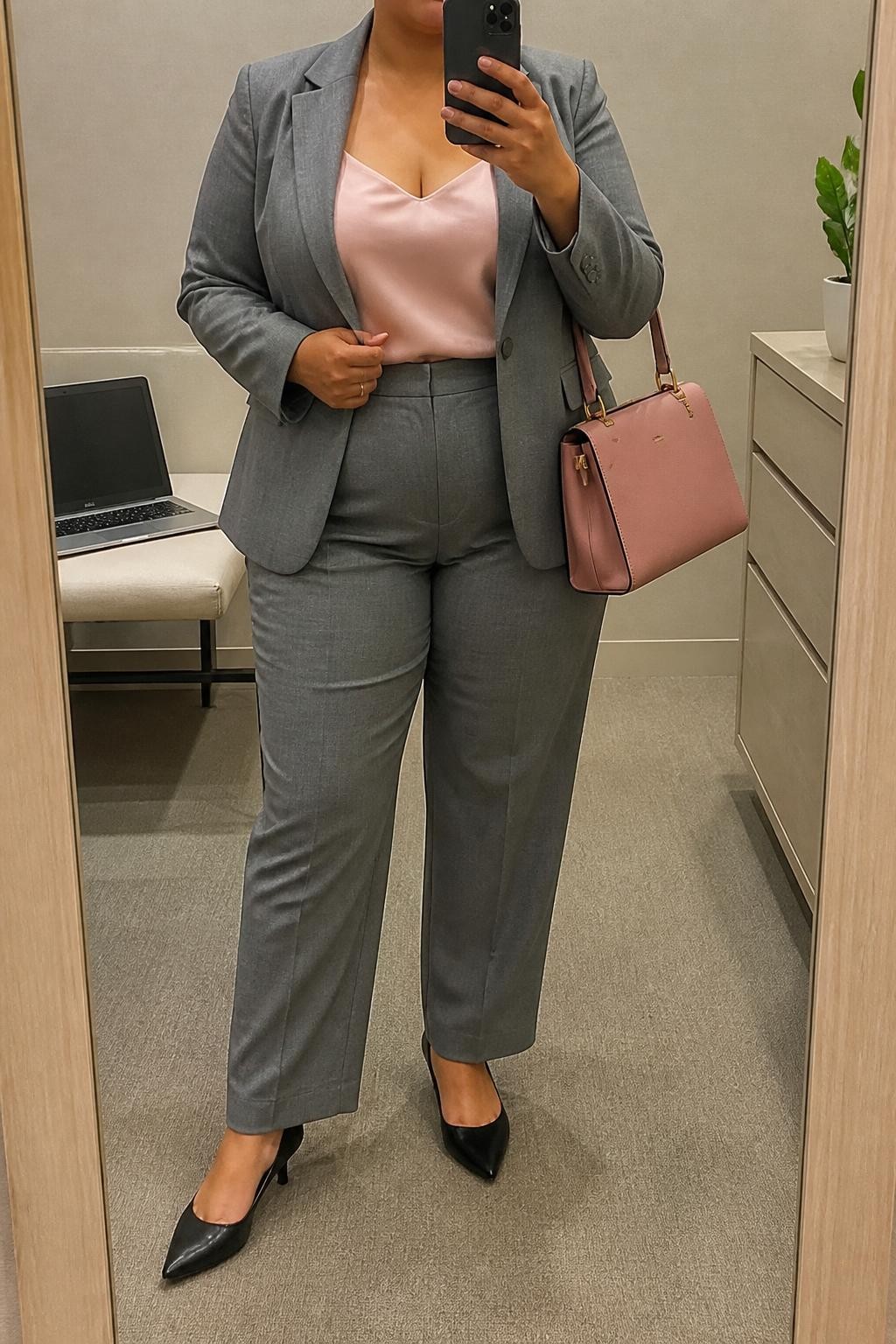Mirror selfie of a plus size woman wearing a medium-grey tailored blazer with matching high-waisted straight-leg trousers, a blush pink satin camisole with wide supportive straps and a modest neckline, black pointed kitten heels, and a structured blush leather handbag, clearly showing the exact suit, grey color, blush satin, footwear, and accessories listed, holding a phone in front of a mirror, cropped at the neck so the face is hidden, relaxed professional selfie stance, in a minimal dressing room with soft indoor lighting, a laptop on a bench and a small plant on a dresser, realistic corporate interview styling, latest iPhone photo quality.