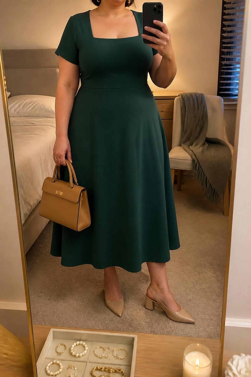 Mirror selfie of a plus size woman wearing a forest green crepe midi dress with short sleeves, a square neckline, a softly fitted waist, and an A-line skirt, nude pointed slingback heels with low block heels, a tan structured handbag, and delicate gold hoop earrings, clearly showing the exact clothing items, green dress, neckline, colors, footwear, and accessories listed, holding a phone in front of a mirror, cropped at the neck so her face does not show, relaxed polished selfie pose, in a cozy bedroom mirror setup with evening golden-hour lighting, a folded blanket on a chair and a jewelry tray on the vanity, realistic interview outfit, latest iPhone photo quality.