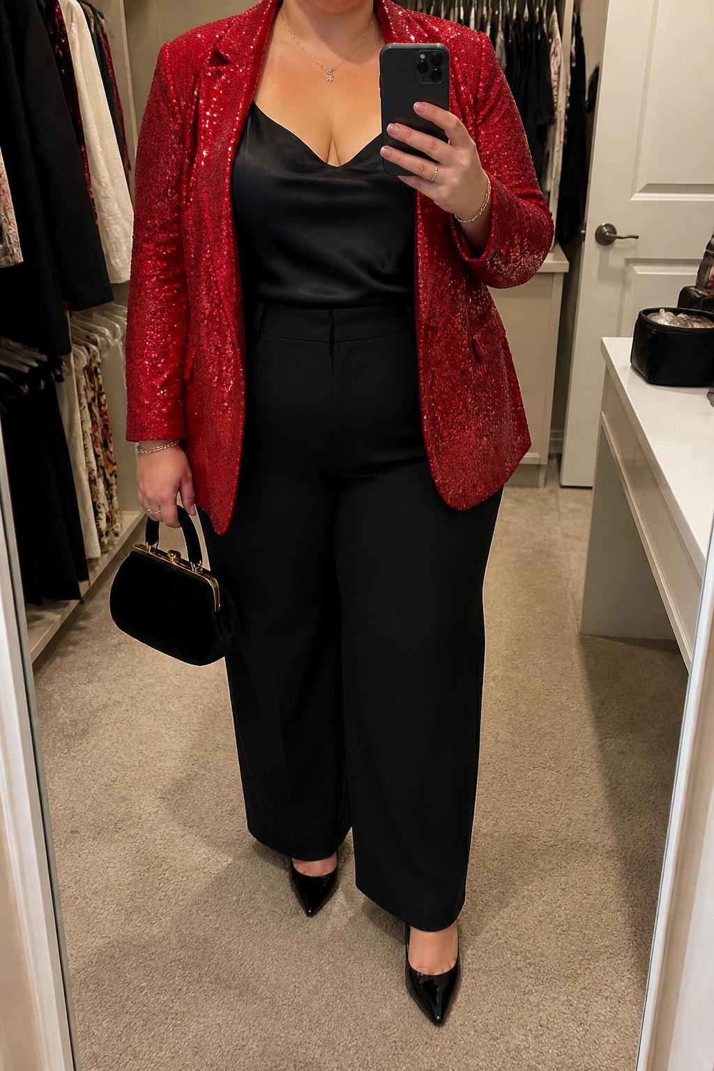 Mirror selfie of a plus size woman wearing a bright red sequin blazer over a fitted black satin camisole, high-waisted black wide-leg trousers, black pointed pumps, and a small black velvet handbag, clearly showing the exact outfit pieces, colors, textures, and accessories listed, holding a phone in front of a mirror, cropped at the neck so her face does not appear, relaxed polished selfie pose, in a walk-in closet mirror with soft indoor lighting, an open closet with clothes visible and a cosmetic bag on the counter, realistic festive evening styling, latest iPhone photo quality.