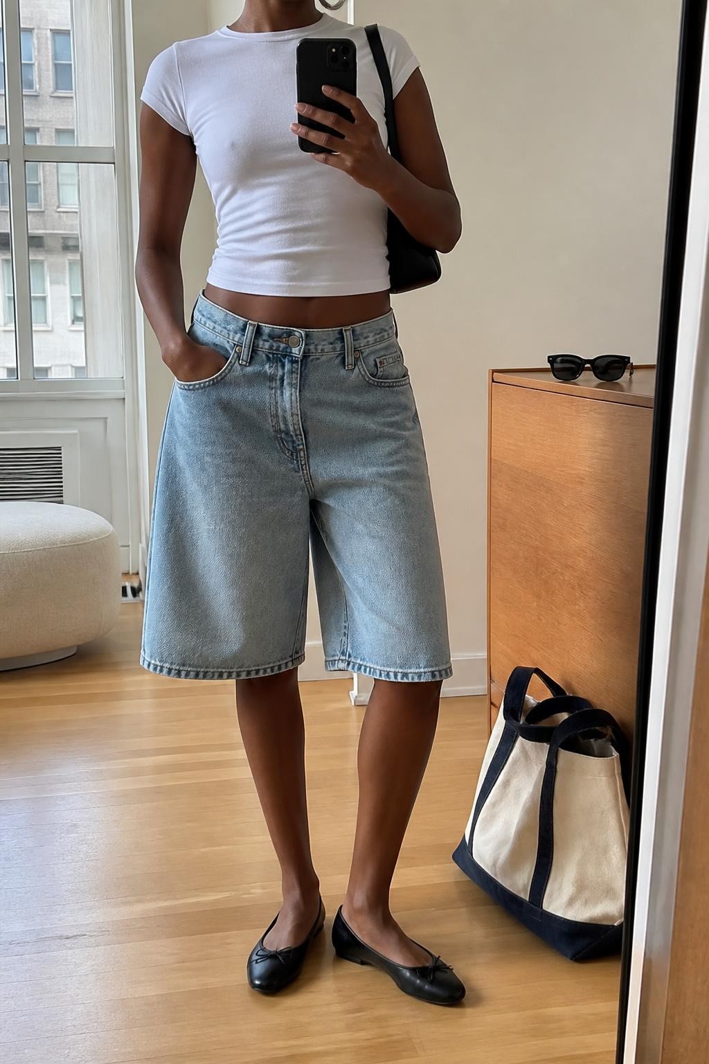 mirror selfie of a Black woman wearing loose knee-length light washed blue denim baggy jorts, a fitted white cotton baby tee, slim black leather ballet flats, a small black leather shoulder bag and medium silver hoop earrings, wearing exactly the clothing and accessories listed with no extra garments, holding a phone in front of a mirror, cropped at NECK so the face does not show, with a relaxed casual selfie pose, in a stylish city apartment, natural window light, sunglasses placed on a dresser and a tote bag resting on the floor, realistic denim folds and natural body proportions, latest iPhone photo quality.