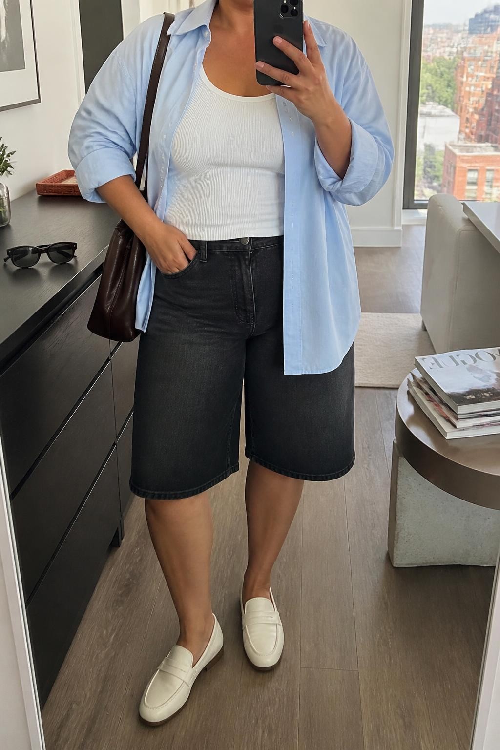 mirror selfie of a plus size woman with warm tan skin wearing long black denim jorts ending near the knee, a pale blue oversized cotton button-up shirt worn open over a fitted white ribbed tank, cream loafers and a dark brown leather shoulder bag, wearing exactly the listed outfit with no additional clothing pieces, holding a phone in front of a mirror, cropped at NECK so the face does not show, relaxed casual selfie pose, in a stylish city apartment, natural window light, sunglasses placed on a dresser and a stack of fashion magazines on a side table, realistic latest iPhone photo quality.
