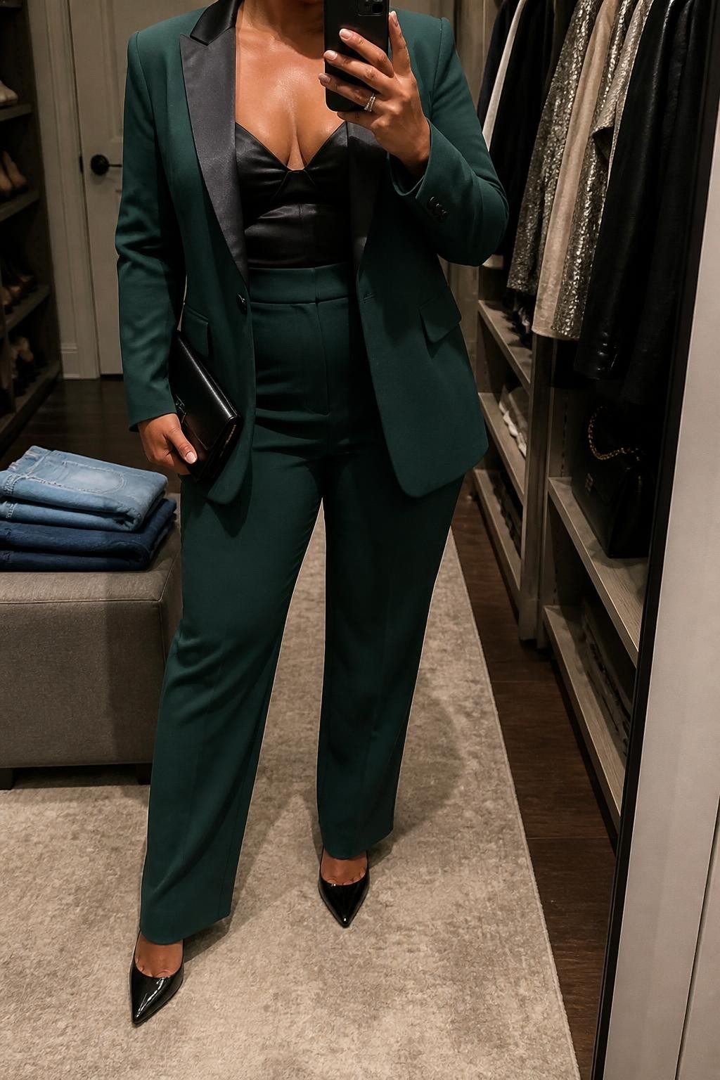 Mirror selfie of a plus size woman wearing a forest green tailored tuxedo blazer with matching high-waisted straight-leg trousers, a black satin bralette-style top with full coverage, black pointed heels, and a slim black clutch, clearly showing the exact outfit pieces, colors, tailoring, fabrics, and accessories listed, holding a phone in front of a mirror, cropped at the neck so her face does not appear, relaxed confident selfie stance, in a walk-in closet mirror with warm moody lighting, folded denim jeans on a bench and a clothing rack with garments behind her, realistic glamorous holiday party appearance, latest iPhone photo quality.