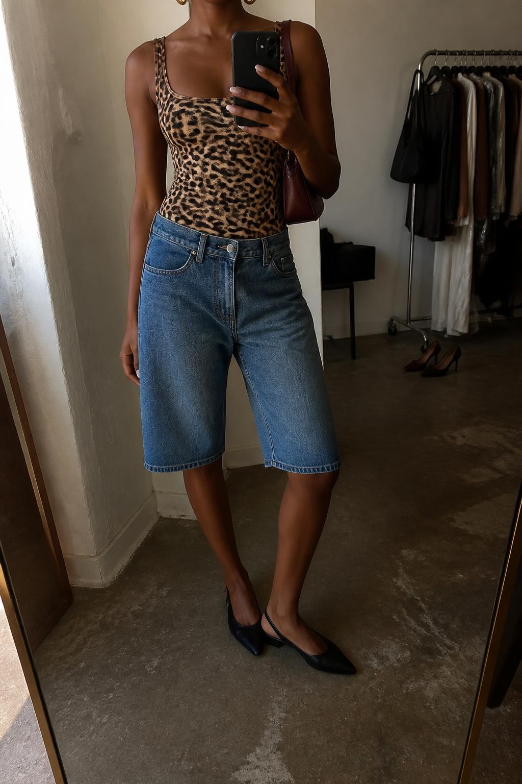 mirror selfie of a Black woman wearing long medium-blue denim jorts with a clean straight cut, a fitted leopard print tank in warm brown and black tones, black leather slingback flats, a small burgundy leather shoulder bag and medium gold hoop earrings, wearing exactly the listed outfit and no additional clothing pieces, holding a phone in front of a mirror, cropped at NECK so the face does not show, relaxed casual selfie pose, in a fashion studio mirror corner, warm moody lighting, clothing rack with garments behind and a pair of heels on the floor nearby, natural unstaged styling, latest iPhone photo quality.