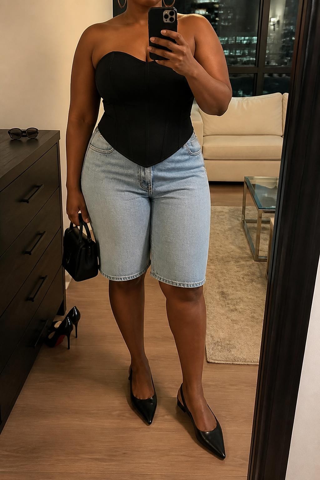 mirror selfie of a plus size Black woman wearing light blue relaxed denim jorts with a knee-length fit, a fitted black cotton corset-inspired top with clean seam detailing, black pointed slingback flats, a structured black mini handbag and medium silver hoop earrings, wearing exactly the listed clothing and accessories with nothing additional, holding a phone in front of a mirror, cropped at NECK so the face does not show, relaxed confident selfie pose, in a stylish city apartment, warm moody lighting, pair of heels on the floor nearby and sunglasses placed on a dresser, realistic polished casual styling, latest iPhone photo quality.