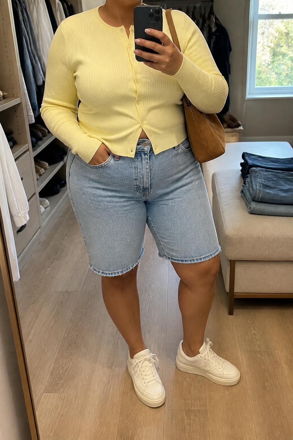 mirror selfie of a plus size woman with medium brown skin wearing light-wash straight-cut denim jorts, a fitted butter yellow fine-knit cardigan buttoned as a top, cream leather sneakers, a tan suede shoulder bag and small gold hoop earrings, wearing exactly the listed clothing and accessories with nothing additional, holding a phone in front of a mirror, cropped at NECK so the face does not show, relaxed casual selfie pose, in a walk-in closet mirror, natural window light, open closet with clothes visible and folded denim jeans on a bench, realistic casual styling, latest iPhone photo quality.
