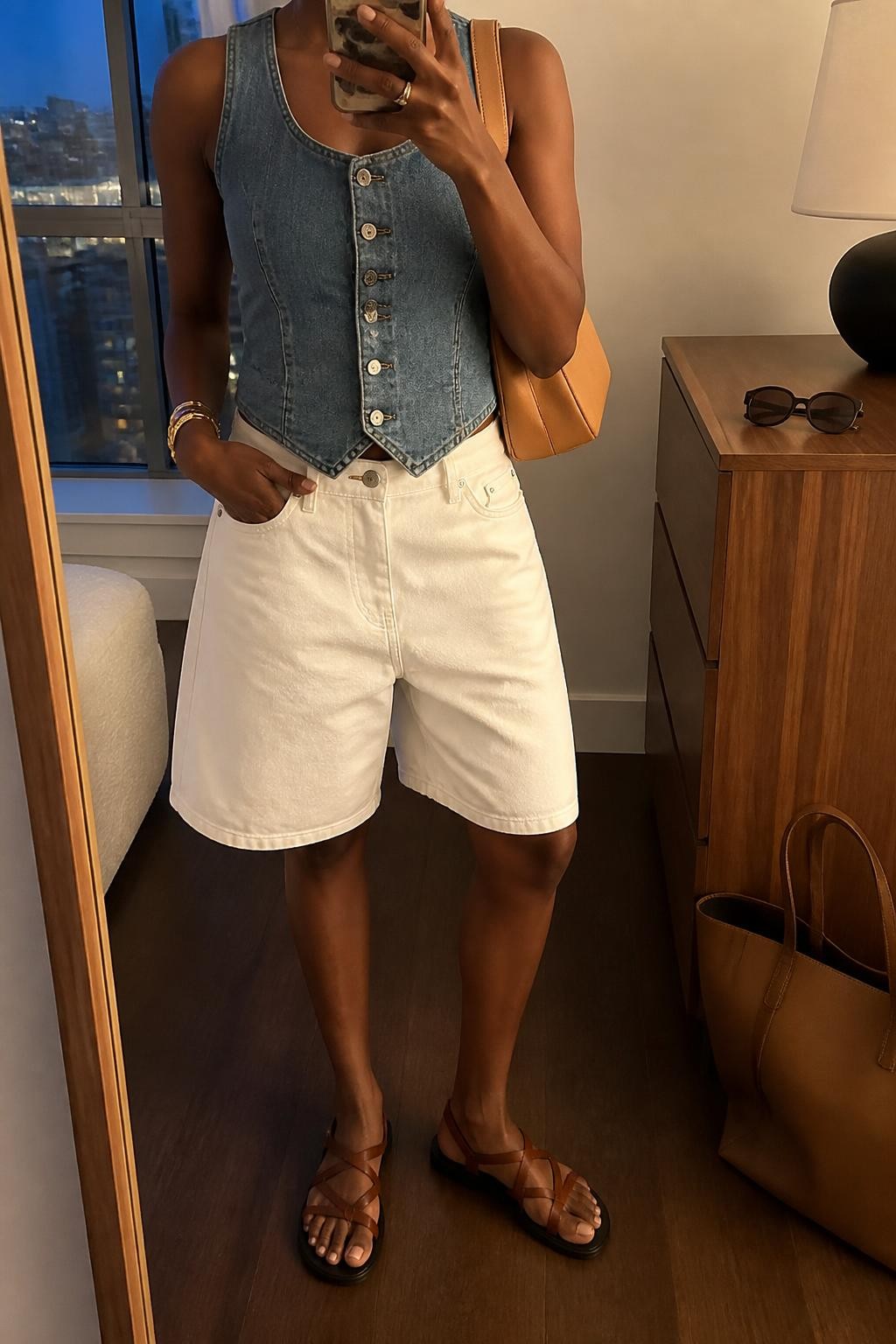 mirror selfie of a Black woman wearing white relaxed denim jorts, a fitted medium-wash blue button-front denim vest, brown leather flat sandals, a tan shoulder bag and small gold hoop earrings, wearing exactly the listed clothing and accessories without extra garments, holding a phone in front of a mirror, cropped at NECK so the face does not show, relaxed casual selfie pose, in a stylish city apartment, evening golden hour, sunglasses placed on a dresser and a tote bag resting on the floor, realistic warm-weather styling, latest iPhone photo quality.