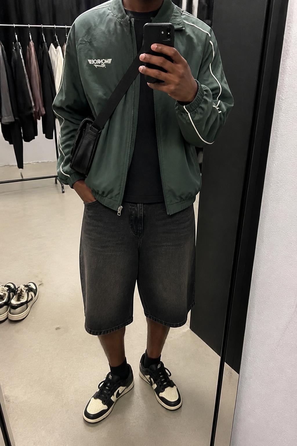 mirror selfie of a Black man wearing washed loose black denim jorts, a forest green lightweight racing-style jacket with subtle cream piping over a plain black cotton T-shirt, black and cream sneakers and a black nylon crossbody bag, wearing the exact pieces listed with no extra clothing, holding a phone in front of a mirror, cropped at NECK so the face does not show, relaxed masculine street-style pose, in a fashion studio mirror corner, soft indoor lighting, clothing rack with garments behind and a pair of sneakers on the floor nearby, realistic unstaged iPhone photography, latest iPhone photo quality.