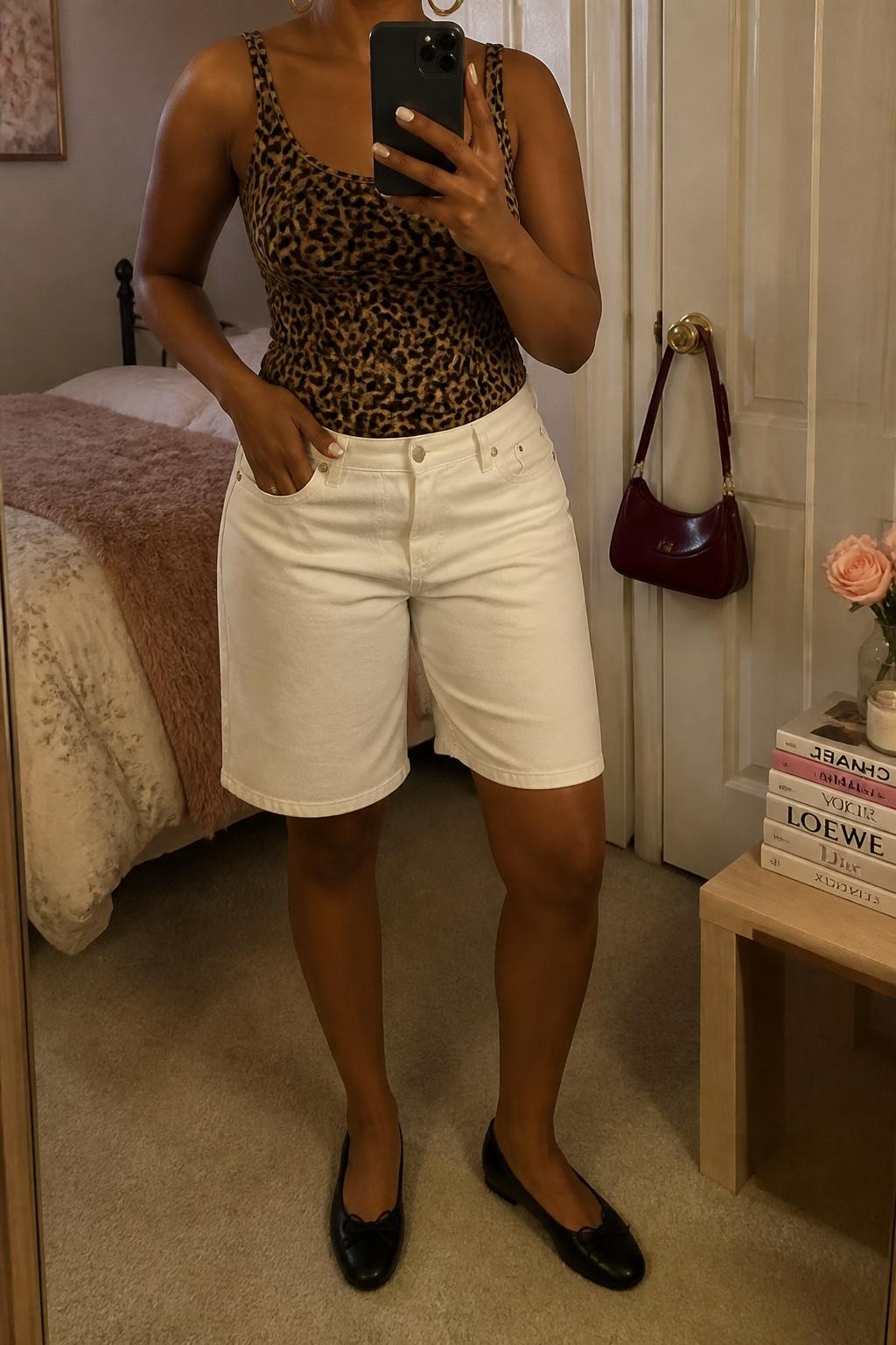 mirror selfie of a Black woman wearing clean white straight-cut denim jorts, a fitted leopard print tank top in warm brown and black tones, black leather ballet flats, a burgundy mini shoulder bag and medium gold hoop earrings, wearing exactly the listed clothing and accessories with no extra garments, holding a phone in front of a mirror, cropped at NECK so the face does not show, relaxed confident selfie pose, in a feminine-themed bedroom, warm moody lighting, handbag hanging on a door handle and a stack of fashion magazines on a side table, realistic casual styling, latest iPhone photo quality.