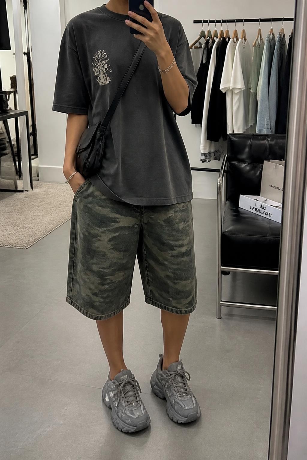 mirror selfie of a woman with medium brown skin wearing baggy green camouflage denim jorts, an oversized charcoal gray cotton graphic T-shirt with a small faded cream graphic, chunky gray sneakers, a black nylon crossbody bag and a silver chain bracelet, wearing exactly the outfit listed with no extra clothing or accessories, holding a phone in front of a mirror, cropped at NECK so the face does not show, relaxed street-style selfie pose, in a fashion studio mirror corner, soft indoor lighting, clothing rack with garments behind and shopping bags beside a chair, realistic casual latest iPhone photo quality.