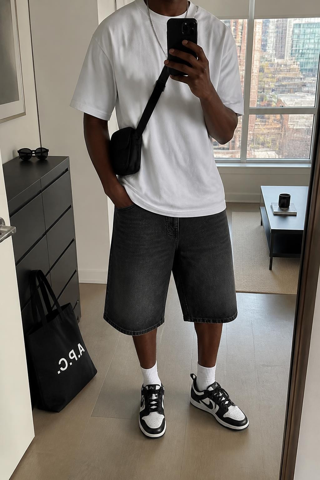 mirror selfie of a Black man wearing baggy knee-length washed black denim jorts, a boxy heavyweight white cotton T-shirt, black and white retro sneakers, a black nylon crossbody bag and a silver chain necklace, wearing exactly the clothing and accessories listed with no extra garments, holding a phone in front of a mirror, cropped at NECK so the face does not show, relaxed masculine casual selfie pose, in a stylish city apartment, natural window light, sunglasses placed on a dresser and a tote bag resting on the floor, realistic fabric folds and natural proportions, latest iPhone photo quality.