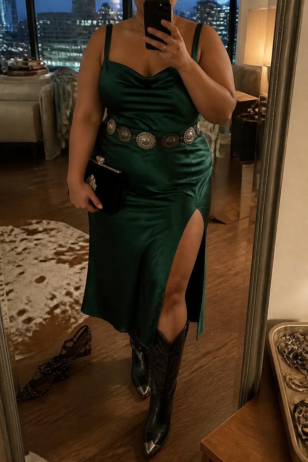 Mirror selfie of a plus size woman wearing an emerald green satin midi dress with wide supportive straps, a softly draped neckline, a fitted waist, and a thigh-high side slit, black leather knee-high Western boots with silver toe details, a black concho belt worn at the waist, and a small black velvet clutch, clearly showing the exact dress, emerald satin, black boots, belt, slit, and accessories listed, holding a phone in front of a mirror, cropped at the neck so her face is hidden, relaxed glamorous selfie pose, in a stylish city apartment with warm moody lighting, a pair of heels on the floor nearby and a jewelry tray on the dresser, realistic vaquera evening outfit, latest iPhone photo quality.