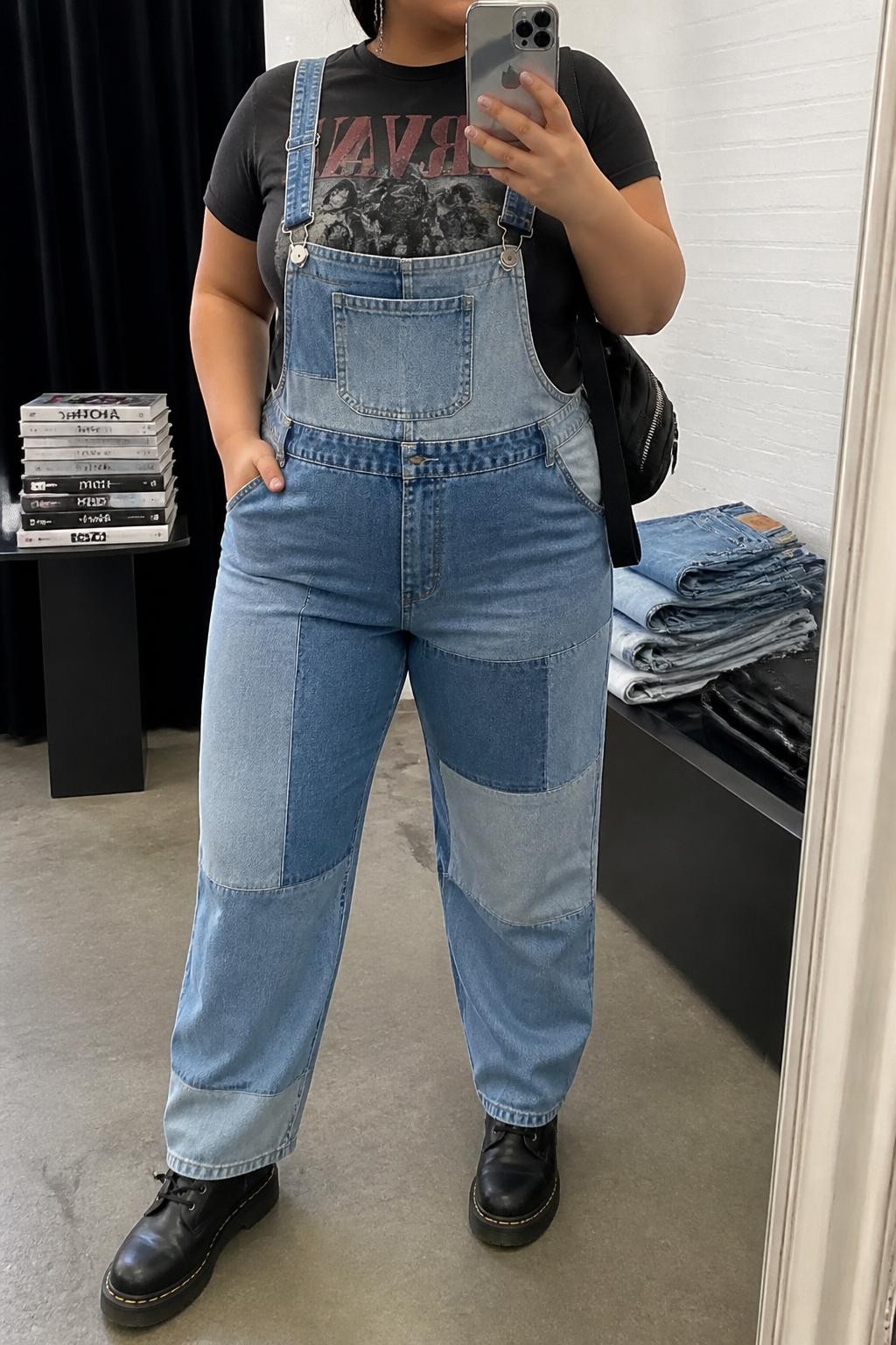 Mirror selfie of a plus size woman wearing patchwork denim overalls with mixed light and medium blue panels, relaxed straight legs, and adjustable straps, layered over a fitted faded black graphic T-shirt, black lace-up combat boots, a compact black nylon backpack, and silver chain earrings, clearly showing the exact clothing items, mixed denim panels, graphic print, footwear, and accessories listed, holding a phone in front of a mirror, cropped at the neck so the face does not show, relaxed edgy selfie pose, in a fashion studio mirror corner with soft indoor lighting, a stack of fashion magazines on a side table and folded denim jeans on a bench, realistic casual concert outfit, latest iPhone photo quality.