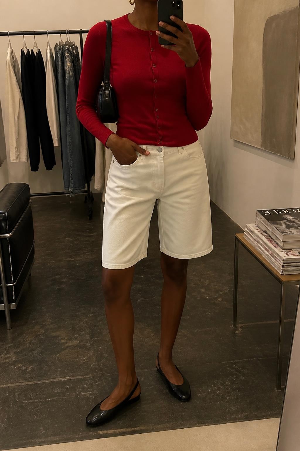 mirror selfie of a Black woman wearing white straight-cut denim jorts, a fitted cherry red fine-knit cardigan buttoned as a top, black leather slingback flats, a compact black shoulder bag and medium gold hoop earrings, wearing exactly the listed clothing and accessories without additional garments, holding a phone in front of a mirror, cropped at NECK so the face does not show, relaxed confident selfie pose, in a fashion studio mirror corner, warm moody lighting, clothing rack with garments behind and a stack of fashion magazines on a side table, believable everyday styling, latest iPhone photo quality.