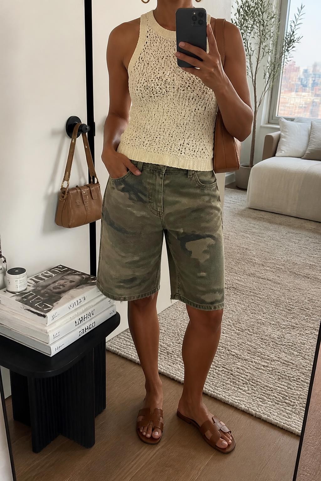 mirror selfie of a woman with medium brown skin wearing faded green and tan camouflage denim jorts with a relaxed knee-length cut, a fitted sleeveless cream crochet top, brown flat leather sandals, a soft tan leather shoulder bag and small gold hoop earrings, wearing exactly the listed clothing and accessories with no extras, holding a phone in front of a mirror, cropped at NECK so the face does not show, relaxed casual selfie pose, in a stylish city apartment, natural window light, handbag hanging on a door handle and a stack of fashion magazines on a side table, realistic warm-weather styling, latest iPhone photo quality.