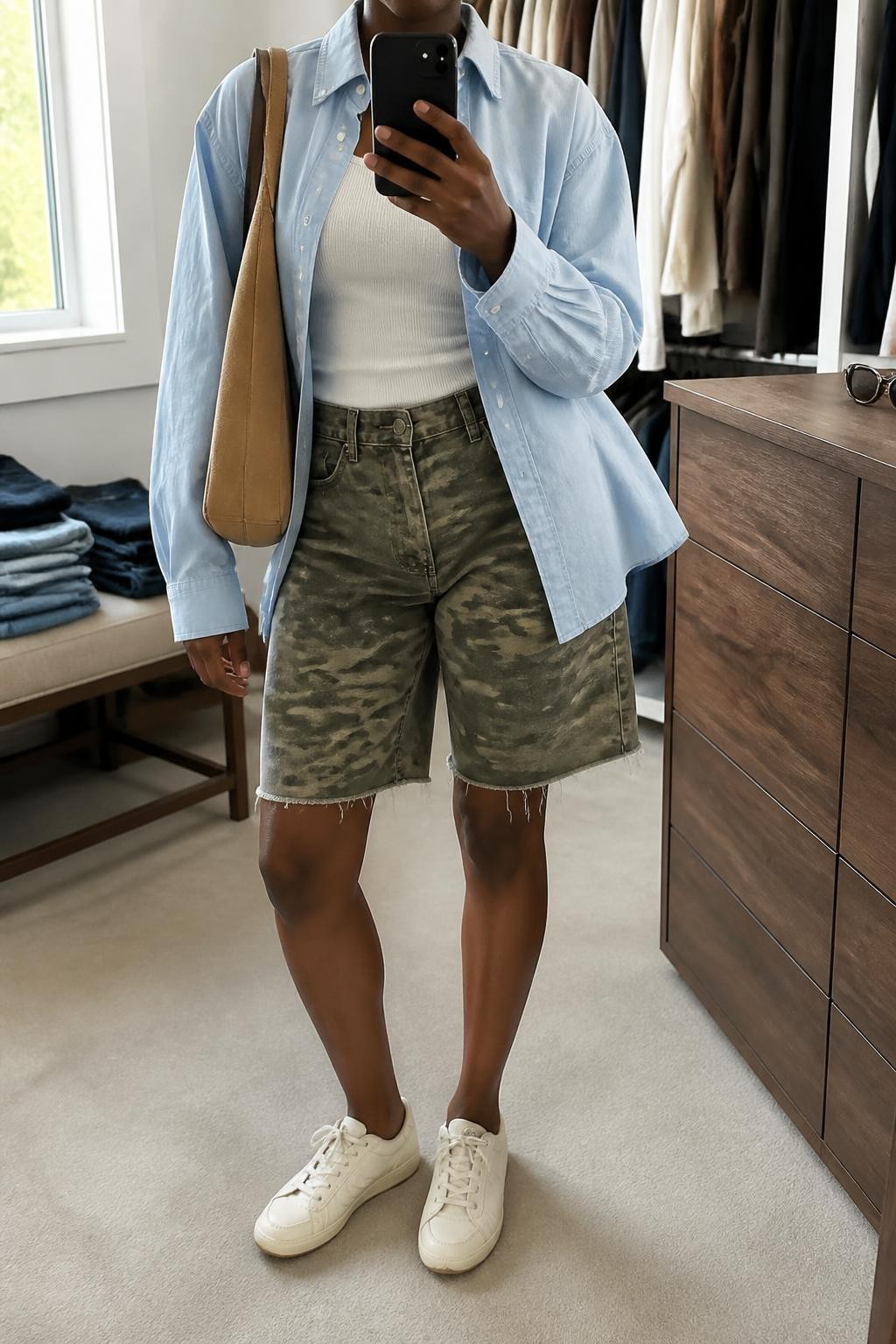 mirror selfie of a Black woman wearing vintage green and brown camouflage denim jorts, a pale blue oversized cotton button-up worn open over a fitted white ribbed tank, cream leather sneakers and a tan suede shoulder bag, wearing exactly the listed outfit with no added garments or accessories, holding a phone in front of a mirror, cropped at NECK so the face does not show, relaxed casual selfie pose, in a walk-in closet mirror, natural window light, folded denim jeans on a bench and sunglasses placed on a dresser, realistic latest iPhone photo quality.