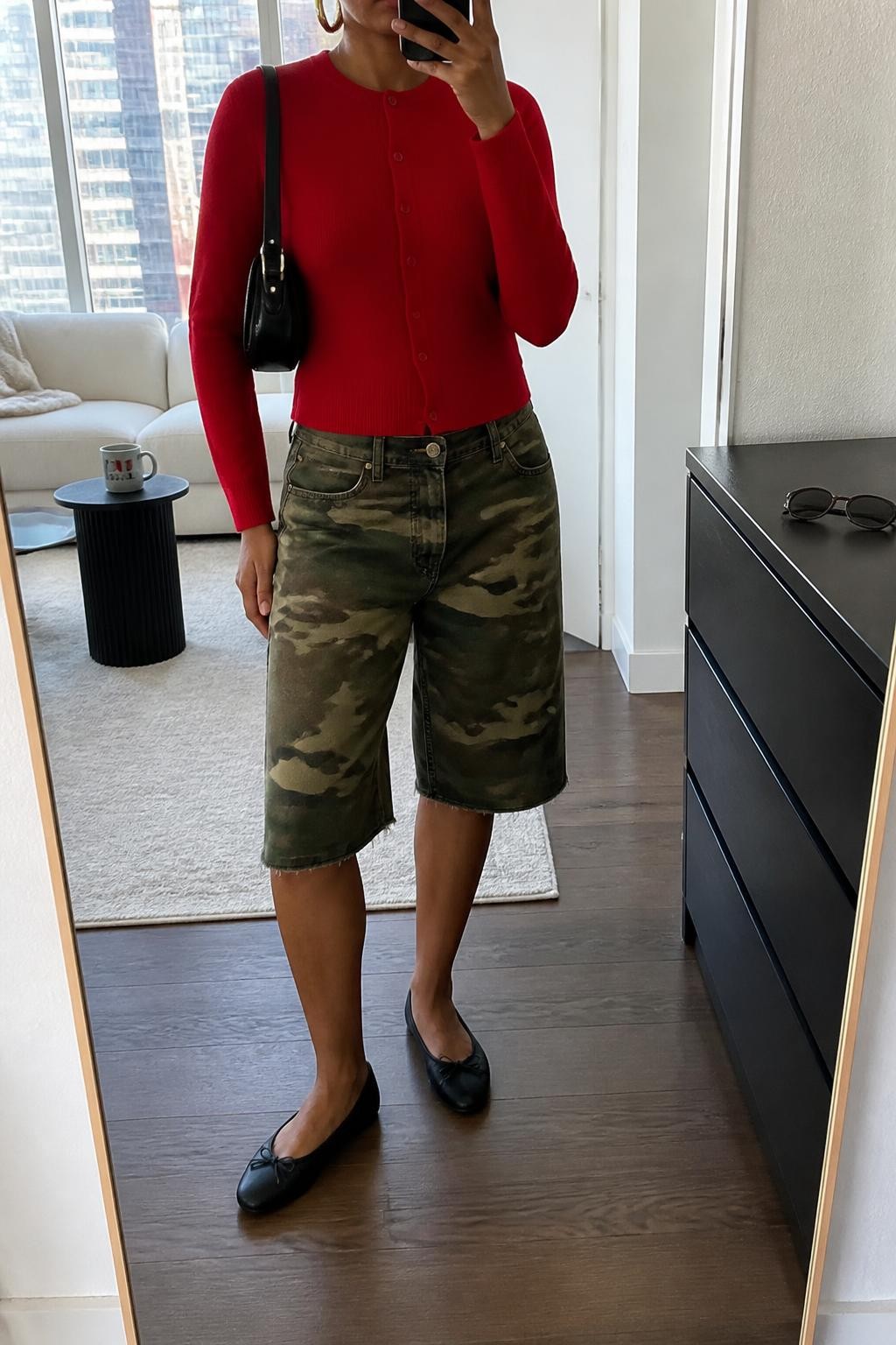 mirror selfie of a Black woman wearing long woodland camo denim jorts ending just below the knee, a fitted cherry red fine-knit cardigan buttoned as a top, black rounded-toe ballet flats, a small black leather shoulder bag and medium gold hoop earrings, wearing exactly the listed clothing and accessories with no added garments, holding a phone in front of a mirror, cropped at NECK so the face does not show, relaxed casual selfie pose, in a stylish city apartment, natural window light, coffee mug on a side table and sunglasses placed on a dresser, natural latest iPhone photo quality.