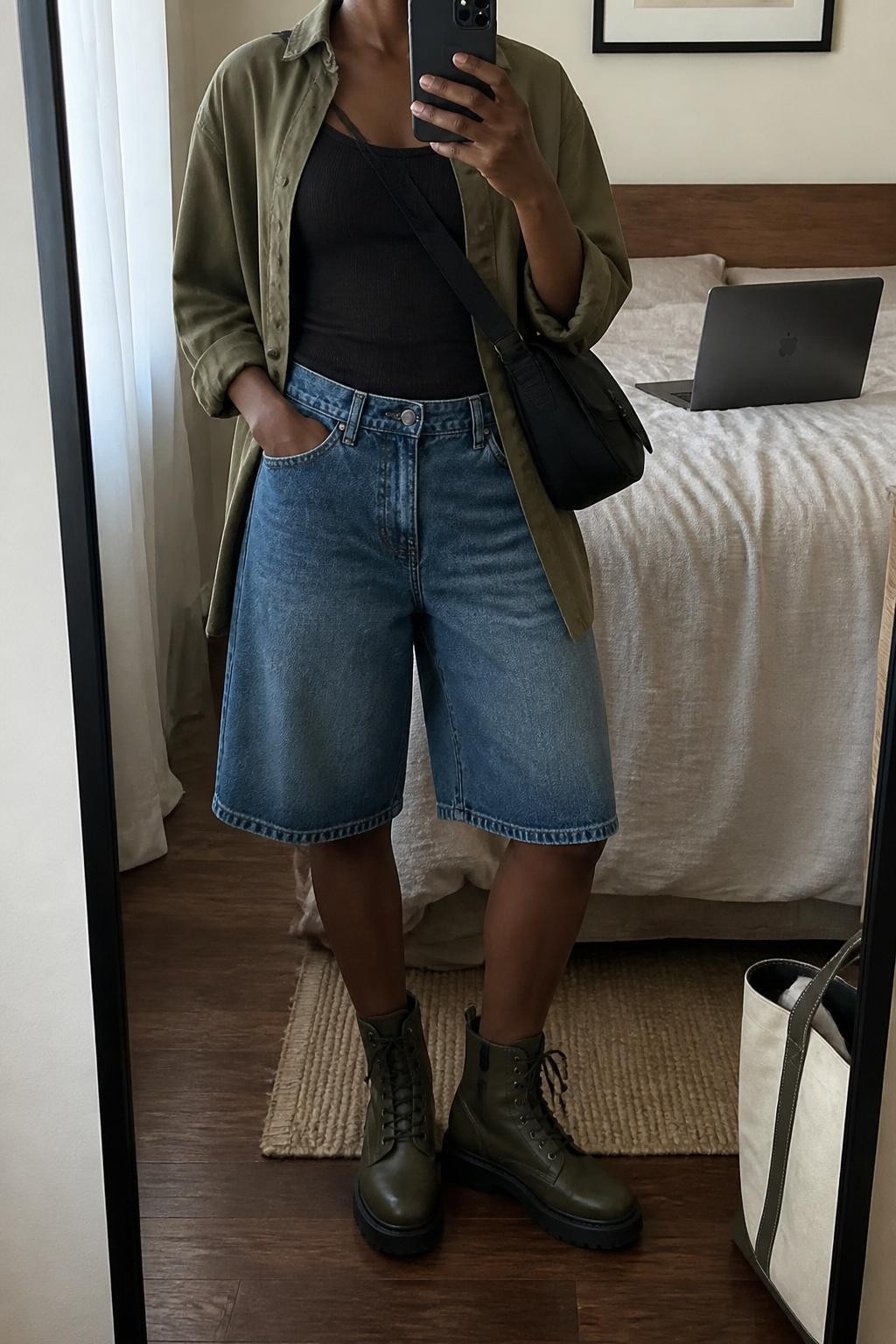 mirror selfie of a Black woman wearing washed blue loose denim jorts, a fitted black ribbed cotton tank layered under an open olive cotton utility shirt, dark olive lace-up utility boots and a black nylon crossbody bag, wearing exactly the listed clothing and accessories with no additional pieces, holding a phone in front of a mirror, cropped at NECK so the face does not show, relaxed casual selfie pose, in a cozy bedroom mirror setup, natural window light, laptop on the bed and a tote bag resting on the floor, realistic casual styling, latest iPhone photo quality.