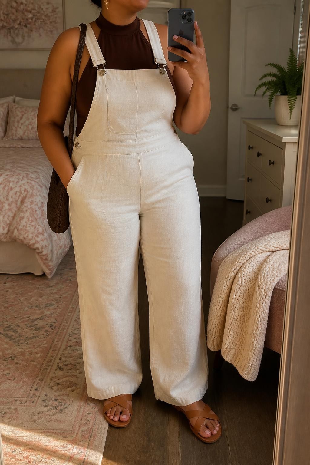 Mirror selfie of a plus size woman wearing cream wide-leg linen overalls with adjustable straps and a softly fitted bib, layered over a chocolate brown halter-neck top with a wide supportive neck band, flat tan leather sandals, a chocolate woven shoulder bag, and delicate gold drop earrings, clearly showing the exact clothing items, cream linen, chocolate top, colors, footwear, and accessories listed, holding a phone in front of a mirror, cropped at the neck so the face does not appear, relaxed polished selfie pose, in a feminine-themed bedroom with evening golden-hour lighting, a folded blanket on a chair and a small plant on a dresser, realistic warm-weather outfit, latest iPhone photo quality.