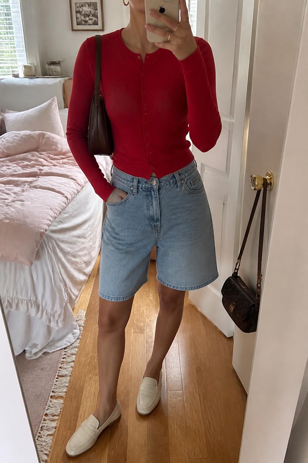 mirror selfie of a woman with light olive skin wearing light blue relaxed denim jorts, a fitted cherry red fine-knit cardigan buttoned as a top, cream leather loafers, a dark brown mini shoulder bag and medium gold hoop earrings, wearing exactly the listed clothing and accessories with no extra garments, holding a phone in front of a mirror, cropped at NECK so the face does not show, relaxed casual selfie pose, in a feminine-themed bedroom, natural window light, soft pillows on an unmade bed and a handbag hanging on a door handle, realistic latest iPhone photo quality.