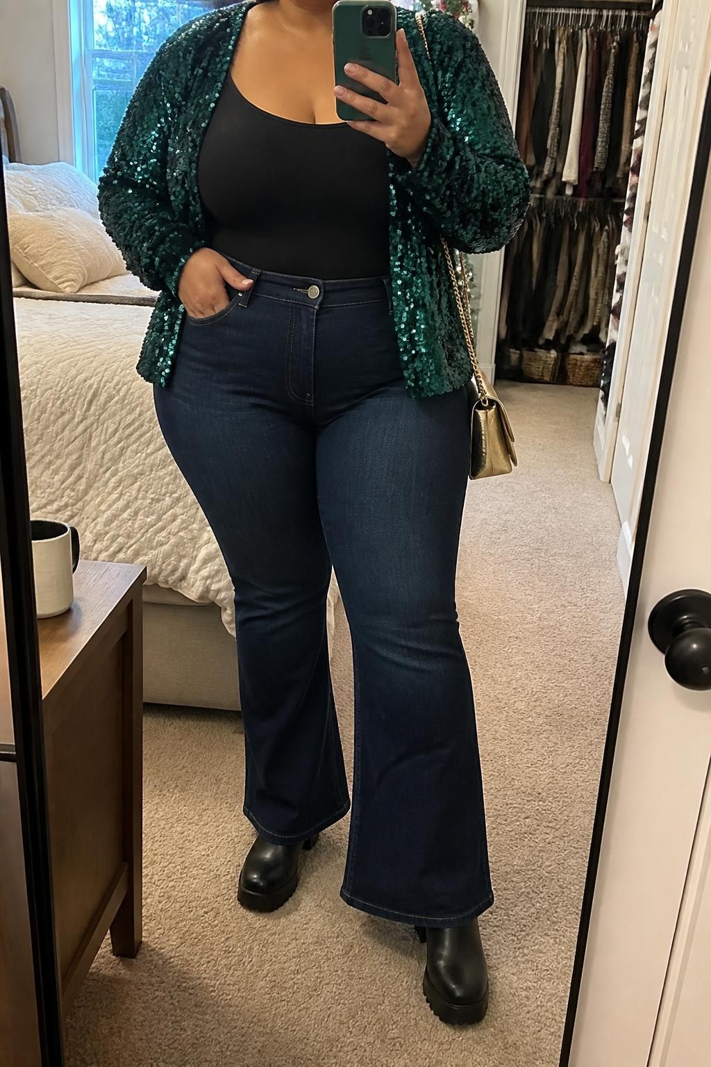 Mirror selfie of a plus size woman wearing dark indigo high-waisted flared jeans, a fitted black scoop-neck bodysuit, an emerald green sequin cardigan worn open, black platform ankle boots, and a small gold shoulder bag, clearly showing the exact clothing items, dark denim, green sequins, colors, fabrics, footwear, and accessories listed, holding a phone in front of a mirror, cropped at the neck so her face does not appear, relaxed casual selfie pose, in a cozy bedroom mirror setup with natural window light, an open closet with clothes visible and a coffee mug on a bedside table, realistic casual Christmas gathering style, latest iPhone photo quality.