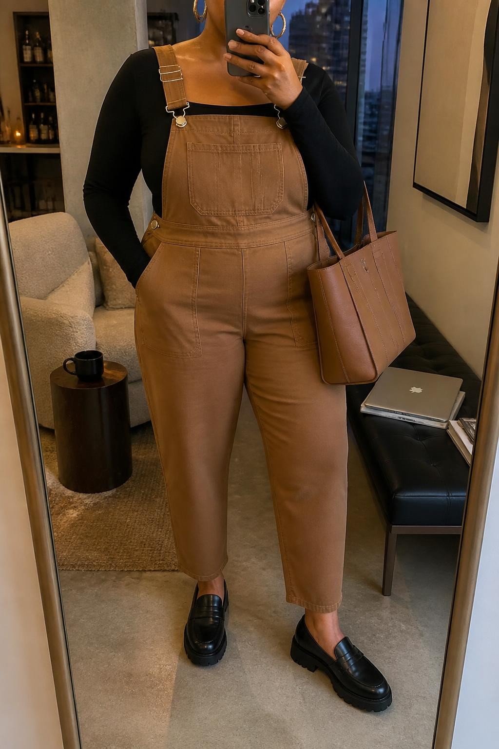 Mirror selfie of a plus size woman wearing camel brown twill overalls with tapered legs, adjustable straps, and flat utility pockets, layered over a fitted black long-sleeve square-neck bodysuit, black leather loafers with chunky soles, a structured camel tote bag, and chunky gold hoop earrings, clearly showing the exact clothing items, camel twill, black fitted top, utility pockets, footwear, and accessories listed, holding a phone in front of a mirror, cropped at the neck so her face is hidden, relaxed polished selfie stance, in a stylish city apartment with warm moody lighting, a coffee mug on a side table and a laptop resting on a bench, realistic everyday outfit, latest iPhone photo quality.