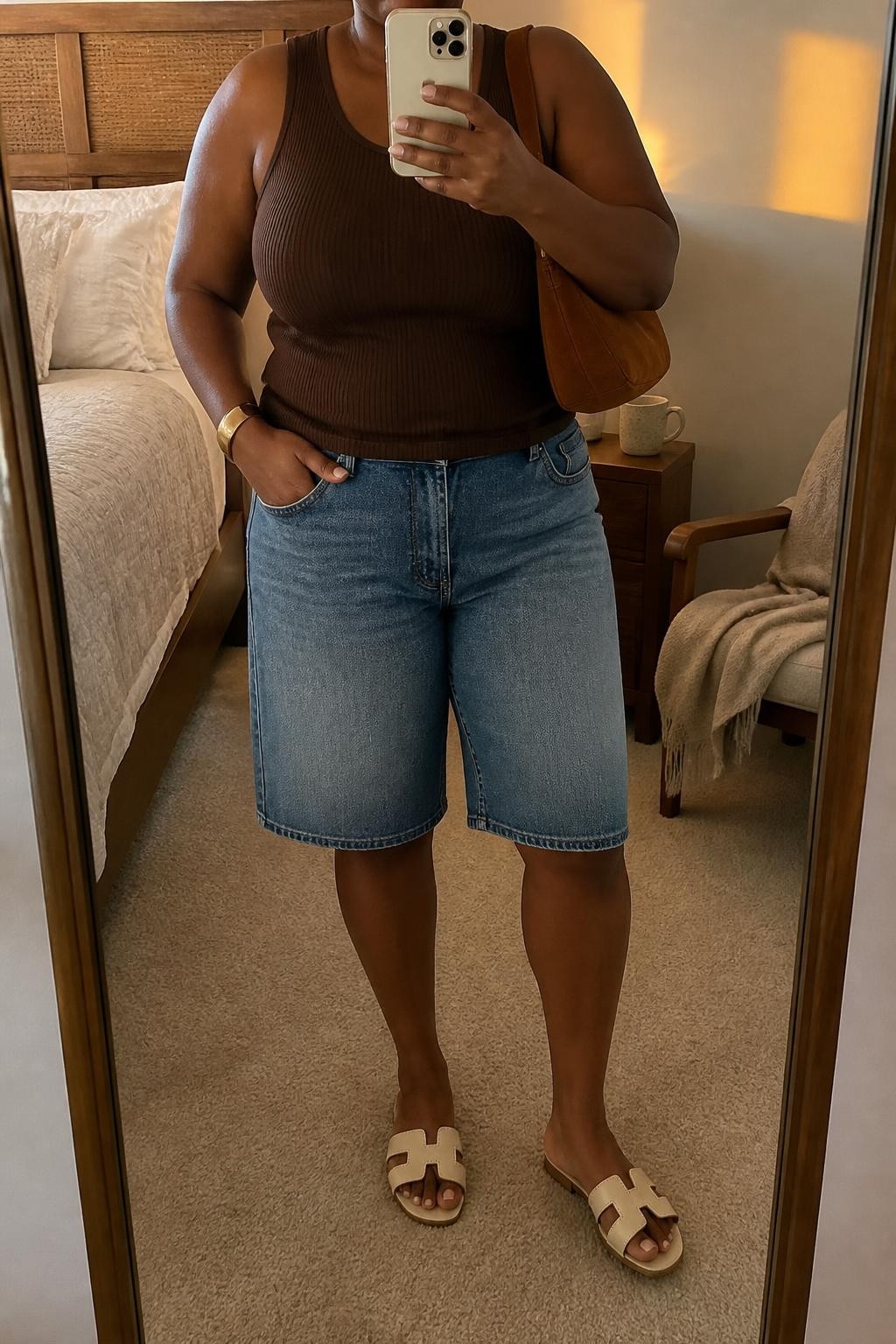 mirror selfie of a plus size Black woman wearing relaxed medium-wash blue denim jorts with a knee-length cut, a fitted chocolate brown ribbed tank top, cream flat leather sandals, a brown suede shoulder bag and a simple gold cuff bracelet, wearing exactly the listed clothing and accessories with no additions, holding a phone in front of a mirror, cropped at NECK so the face does not show, relaxed casual selfie pose, in a cozy bedroom mirror setup, evening golden hour, coffee mug on a bedside table and a folded blanket on a chair, natural warm-weather styling, latest iPhone photo quality.