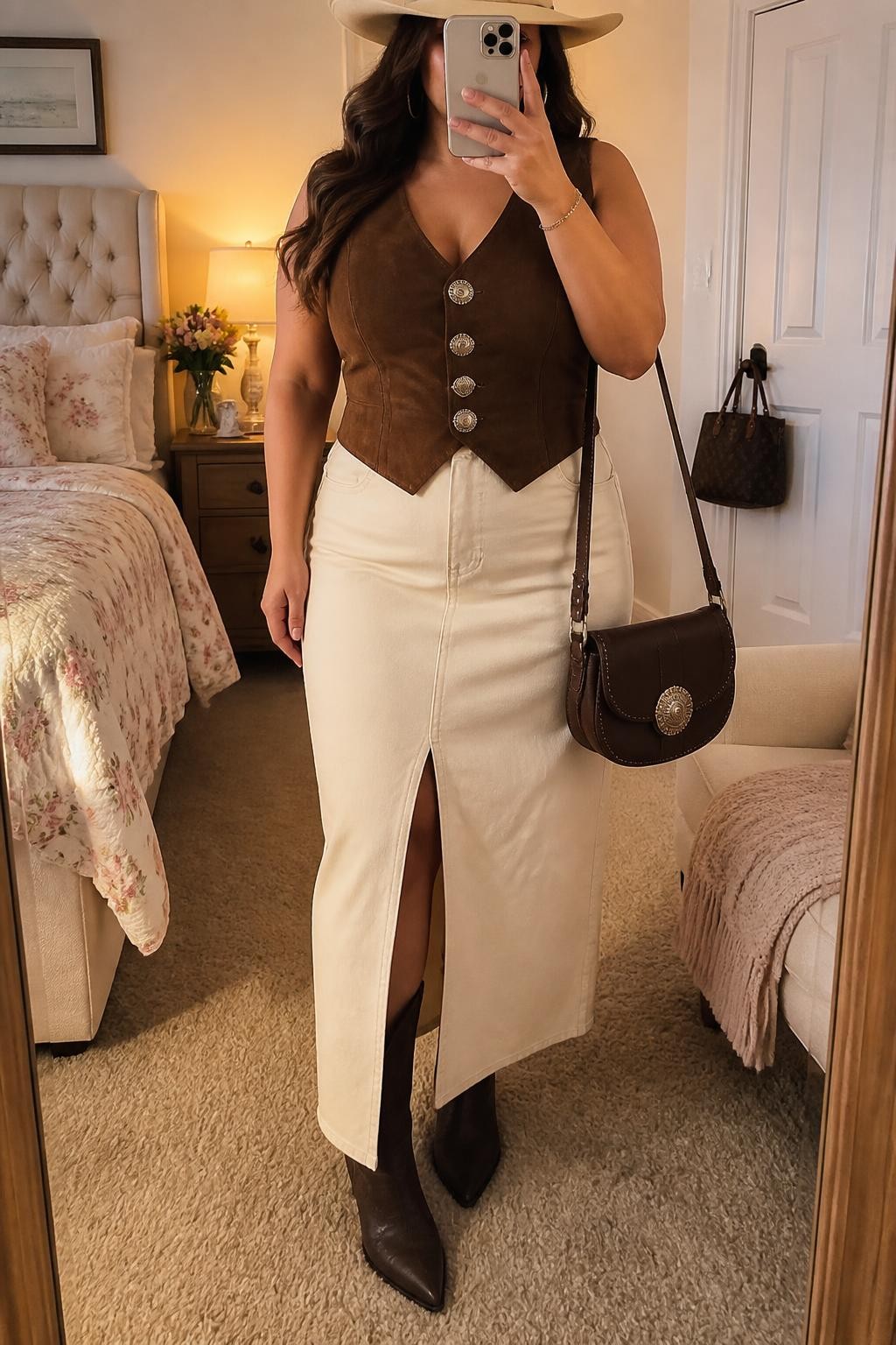 Mirror selfie of a plus size woman wearing a fitted chocolate brown suede Western vest with silver concho buttons, a cream high-waisted denim maxi skirt with a front walking slit, chocolate brown pointed ankle cowboy boots, a cream cowboy hat, and a structured chocolate brown saddle bag, clearly showing the exact clothing items, suede texture, cream denim, concho buttons, footwear, hat, and accessories listed, holding a phone in front of a mirror, cropped at the neck so her face does not show, relaxed polished selfie pose, in a feminine-themed bedroom with evening golden-hour lighting, a folded blanket on a chair and a handbag hanging on a door handle, realistic neutral vaquera outfit, latest iPhone photo quality.