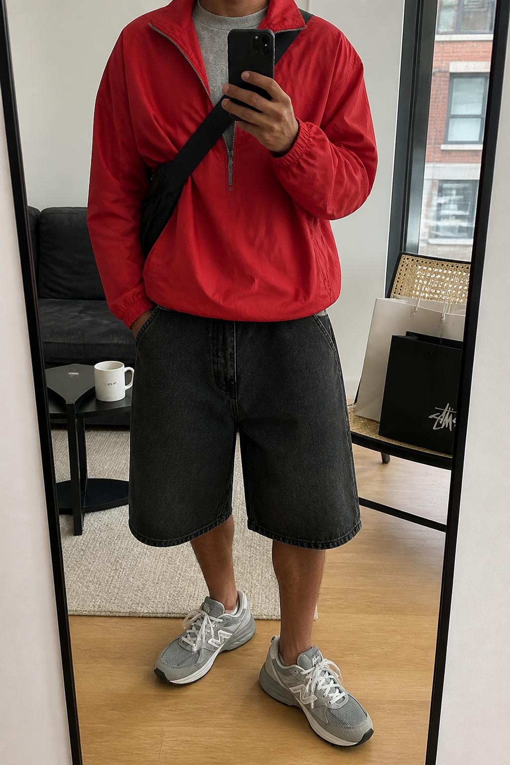 mirror selfie of a man with medium brown skin wearing baggy washed black denim jorts, a lightweight bright red nylon windbreaker zipped halfway over a plain gray cotton T-shirt, gray running sneakers and a black nylon sling bag, wearing the exact listed outfit with no added clothing pieces or accessories, holding a phone in front of a mirror, cropped at NECK so the face does not show, relaxed streetwear selfie pose, in a stylish city apartment, soft indoor lighting, shopping bags beside a chair and a coffee mug on a side table, natural latest iPhone photo quality.