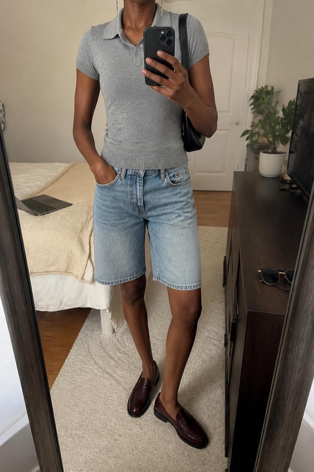 mirror selfie of a Black woman wearing faded blue straight-cut denim jorts, a fitted heather gray short-sleeve fine-knit polo, burgundy leather loafers, a black mini shoulder bag and slim silver drop earrings, wearing exactly the listed outfit without additional garments or accessories, holding a phone in front of a mirror, cropped at NECK so the face does not show, relaxed polished selfie pose, in a cozy bedroom mirror setup, soft indoor lighting, laptop on the bed and sunglasses placed on a dresser, natural latest iPhone photo quality.