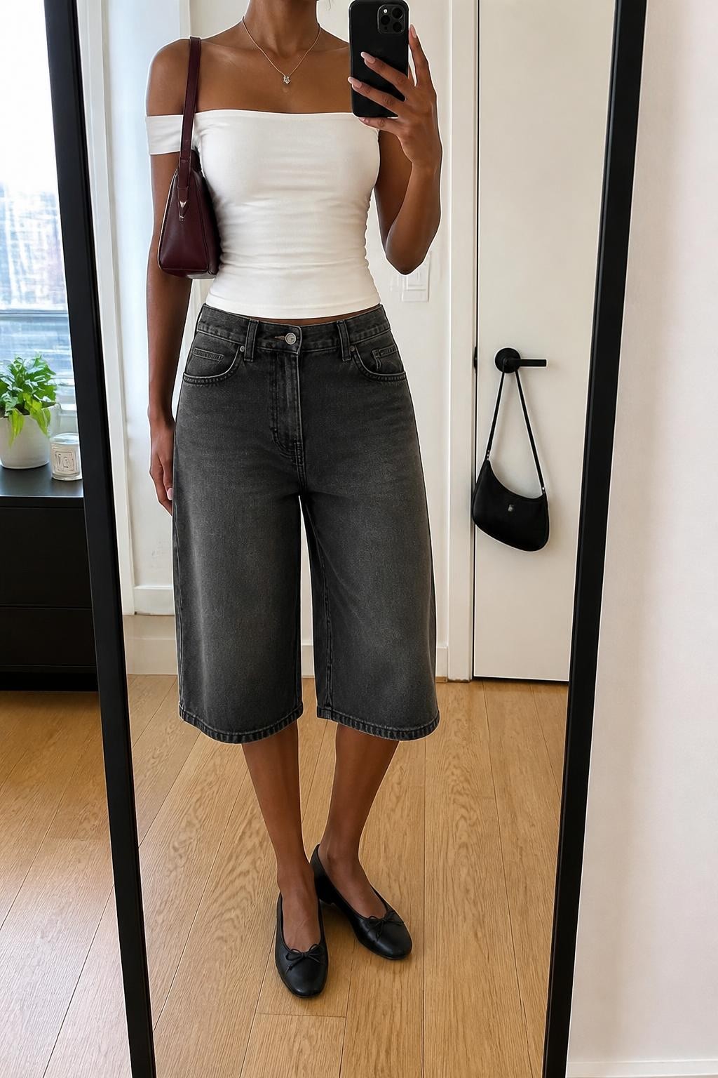 mirror selfie of a Black woman wearing long charcoal gray denim jorts with a relaxed straight cut, a fitted white off-shoulder stretch cotton top, black leather ballet flats, a burgundy mini shoulder bag and a thin silver pendant necklace, wearing exactly the listed outfit with no extra garments, holding a phone in front of a mirror, cropped at NECK so the face does not show, relaxed casual selfie pose, in a stylish city apartment, natural window light, a small plant on a dresser and a handbag hanging on a door handle, realistic latest iPhone photo quality.