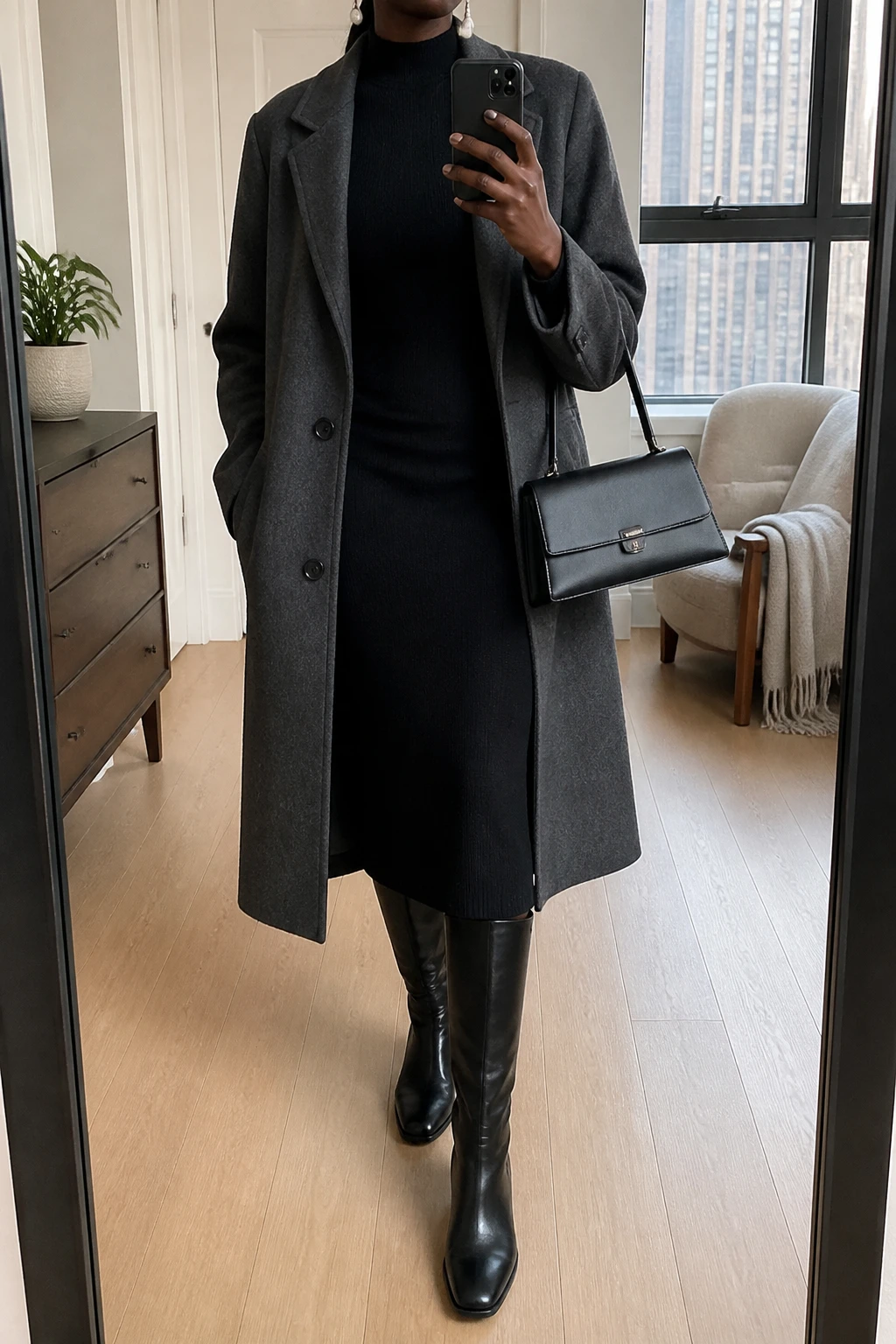 Image Prompt: Mirror selfie of a black woman wearing a long charcoal grey single-breasted wool coat over a black long-sleeve fine-knit cashmere midi dress with a high neckline and gently fitted waist, black polished leather knee-high boots with low block heels, a structured black leather top-handle handbag, and classic white pearl drop earrings, holding a phone in front of a mirror, cropped at the NECK so the face does not show, with a relaxed casual selfie pose, in a stylish city apartment, soft natural window light, a small plant on a dresser and a folded blanket on a chair, realistic black knit fabric, soft charcoal wool coat folds, natural elegant proportions, latest iPhone photo quality, no additional clothing or accessories.