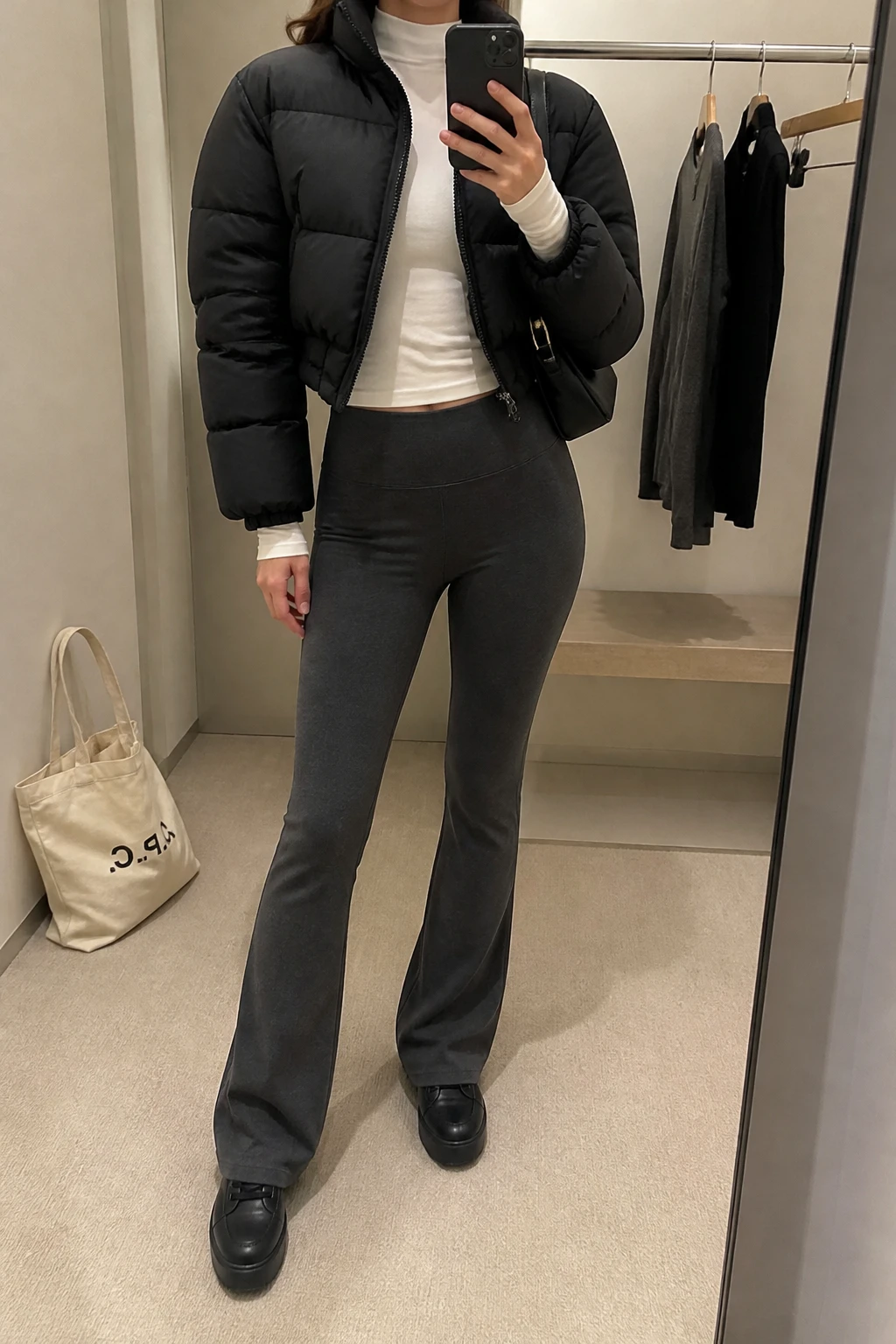 Image Prompt: Mirror selfie of a woman wearing a black cropped insulated quilted puffer jacket over a white fitted long-sleeve thermal mock-neck top, charcoal grey high-waisted fleece-lined flared leggings, black chunky leather sneakers with thick flat rubber soles, and a small black nylon shoulder bag, holding a phone in front of a mirror, cropped at the NECK so the face does not show, with a relaxed casual selfie pose, in a minimal dressing room, soft indoor lighting, a tote bag resting on the floor and a clothing rack with garments behind, realistic black puffer volume, natural charcoal stretch fabric and sporty casual styling, latest iPhone photo quality, no additional clothing or accessories.