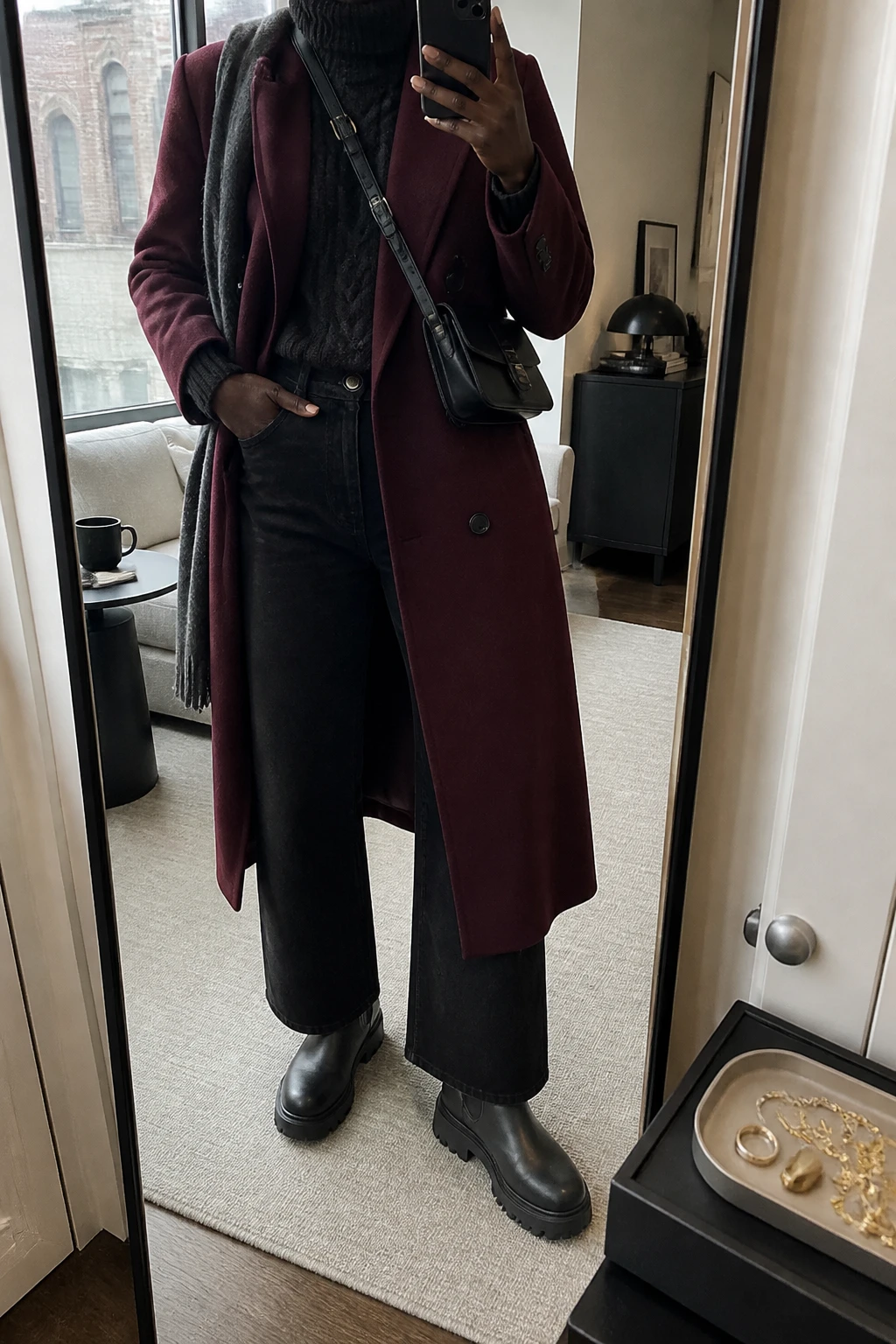 Image Prompt: Mirror selfie of a black woman wearing a long deep burgundy heavyweight double-breasted wool coat over a black chunky cable-knit turtleneck sweater, solid black high-waisted wide-leg denim jeans with black thermal leggings underneath, black waterproof insulated chunky Chelsea boots with flat rubber lug soles, a black ribbed knit beanie, a charcoal grey wool scarf, and a small black leather crossbody bag, holding a phone in front of a mirror, cropped at the NECK so the face does not show, with a relaxed casual selfie pose, in a stylish city apartment, soft natural window light, a coffee mug on a side table and a jewelry tray on the vanity, realistic deep burgundy wool texture, natural black chunky-knit stitches and straight denim creases, latest iPhone photo quality, no additional clothing or accessories.