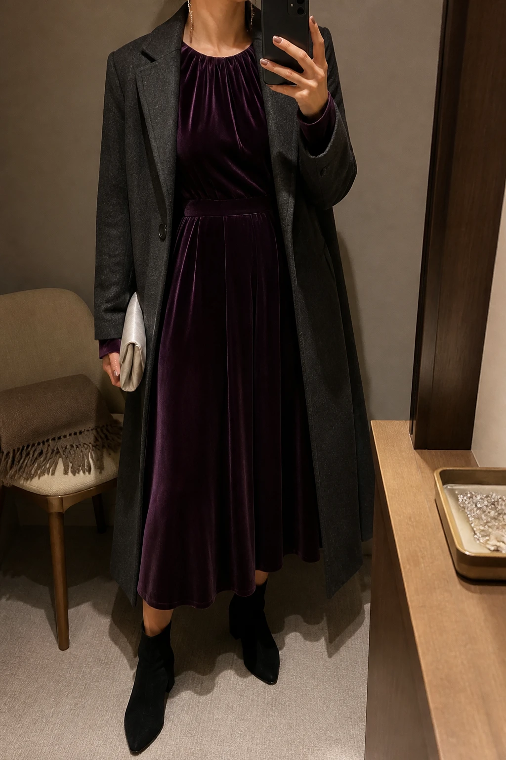 Image Prompt: Mirror selfie of a woman wearing a deep plum long-sleeve velvet midi dress with a softly gathered neckline, gently defined waist, and flowing skirt, black suede pointed-toe ankle boots with low block heels, a long charcoal grey tailored wool coat worn open, a small silver satin clutch, and delicate silver crystal drop earrings, holding a phone in front of a mirror, cropped at the NECK so the face does not show, with a relaxed casual selfie pose, in a minimal dressing room, warm moody indoor lighting, a folded blanket on a chair and a jewelry tray on the vanity, realistic plum velvet texture, natural charcoal wool folds, soft evening shadows, latest iPhone photo quality, no additional clothing or accessories.