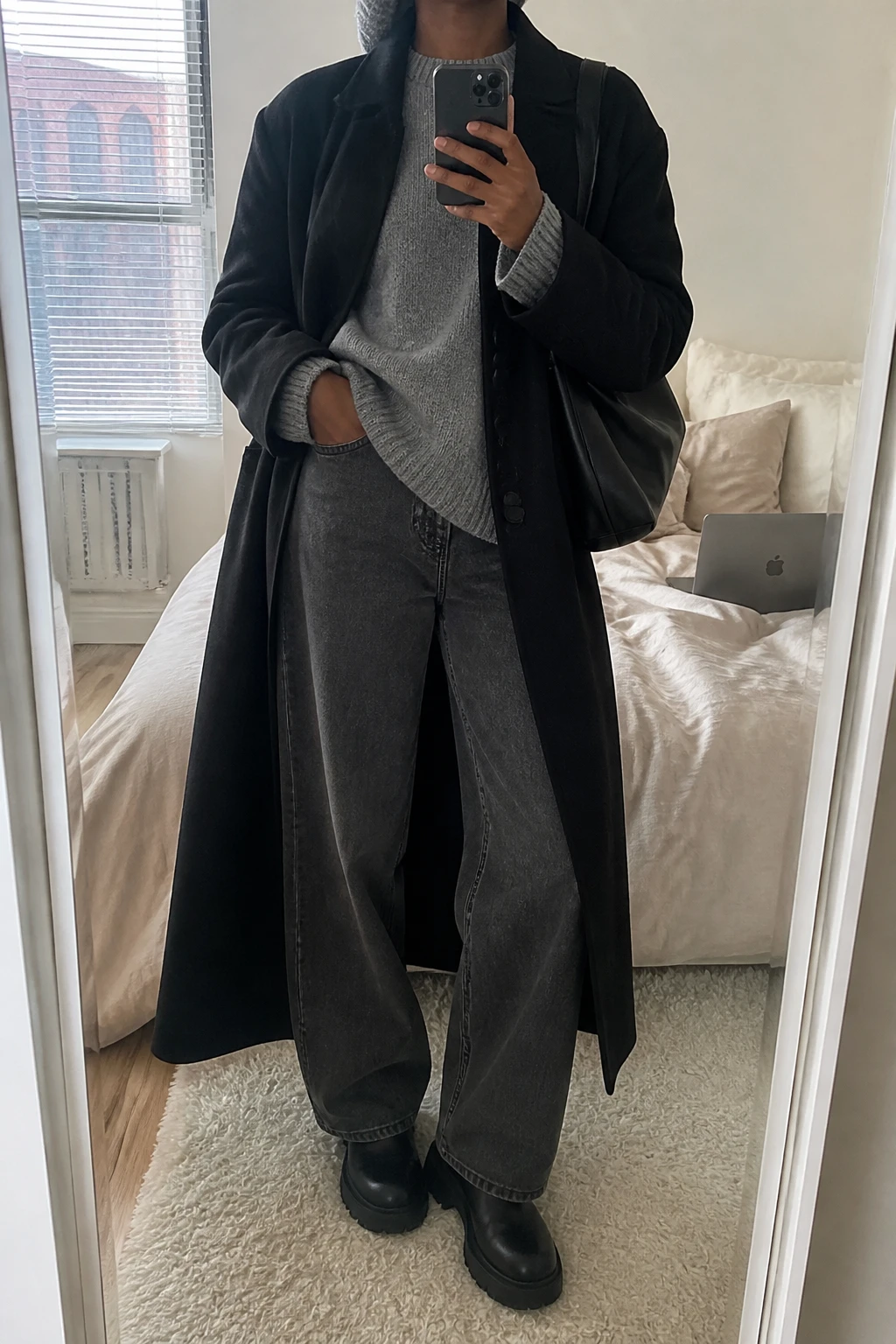 Image Prompt: Mirror selfie of a black woman wearing a long black single-breasted wool maxi coat open over a heather grey chunky crewneck sweater, charcoal grey wide-leg denim jeans, black leather waterproof chunky Chelsea boots, a black leather slouchy shoulder bag, and a grey ribbed knit beanie, holding a phone in front of a mirror, cropped at the NECK so the face does not show, with a relaxed casual selfie pose, in a cozy bedroom mirror setup, soft natural window light, soft pillows on an unmade bed and a laptop on the bed, realistic long coat drape, natural denim folds, latest iPhone photo quality, no additional clothing or accessories.