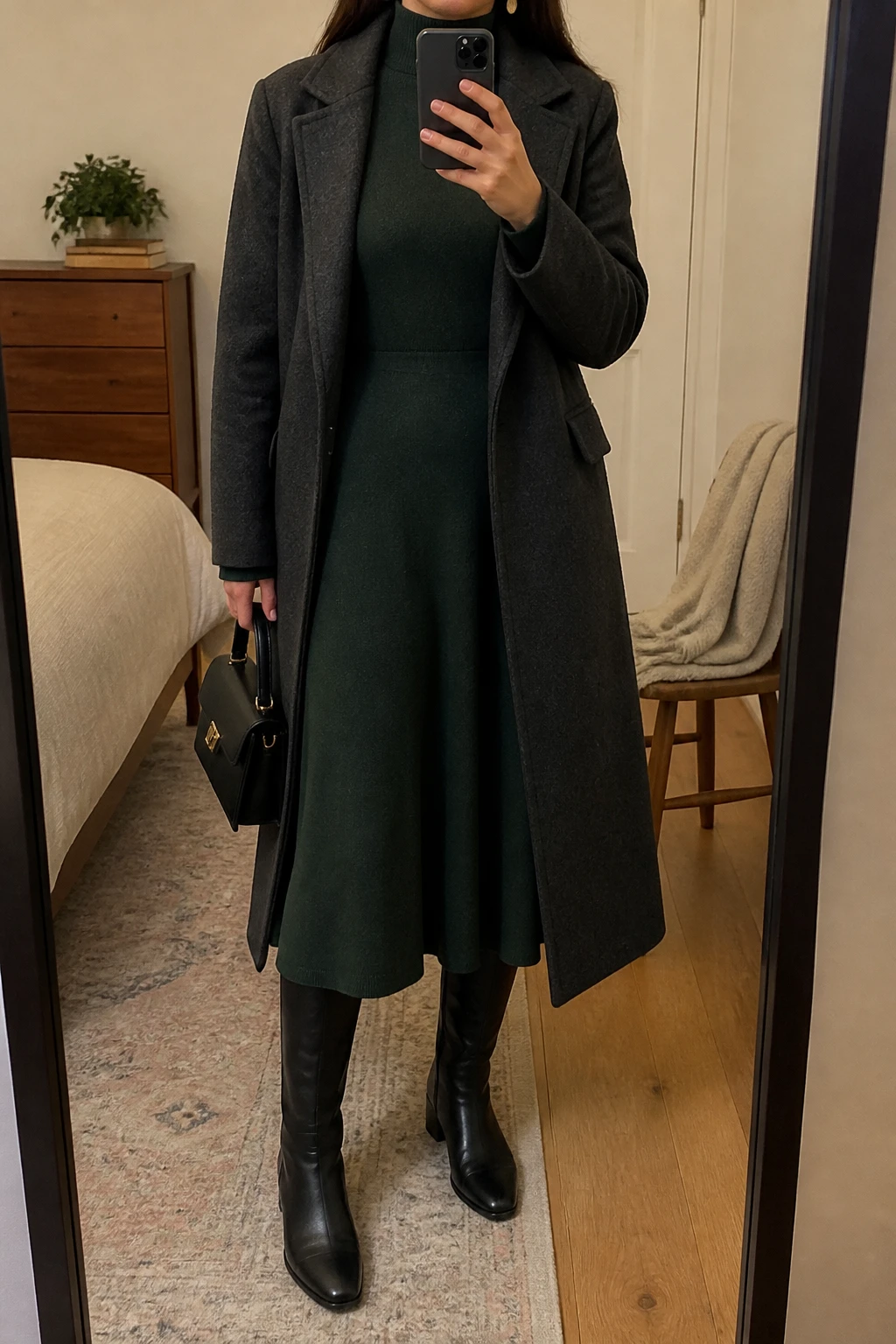 Image Prompt: Mirror selfie of a woman wearing a forest green long-sleeve fine-knit midi dress with a high neckline and gently flared skirt, black leather knee-high boots with low block heels, a long charcoal grey tailored wool coat worn open, a small structured black leather handbag, and delicate gold oval earrings, holding a phone in front of a mirror, cropped at the NECK so the face does not show, with a relaxed casual selfie pose, in a cozy bedroom mirror setup, warm soft indoor lighting, a folded blanket on a chair and a small plant on a dresser, realistic forest green knit fabric, natural flared dress folds and charcoal wool coat texture, latest iPhone photo quality, no additional clothing or accessories.