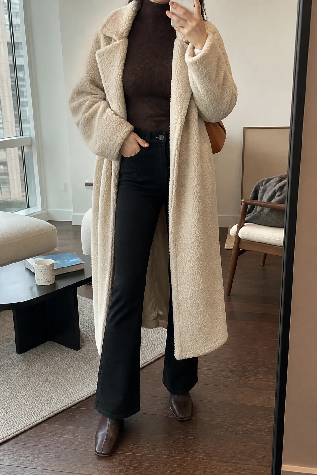 Image Prompt: Mirror selfie of a woman wearing a long cream oversized teddy-fleece coat over a chocolate brown fitted cashmere turtleneck sweater, black high-waisted flared denim jeans, dark brown leather square-toe ankle boots with low block heels, and a small caramel leather shoulder bag, holding a phone in front of a mirror, cropped at the NECK so the face does not show, with a relaxed casual selfie pose, in a stylish city apartment, soft natural window light, a coffee mug on a side table and a folded blanket on a chair, realistic cream teddy-fleece texture, natural chocolate cashmere knit and black flared denim folds, latest iPhone photo quality, no additional clothing or accessories.