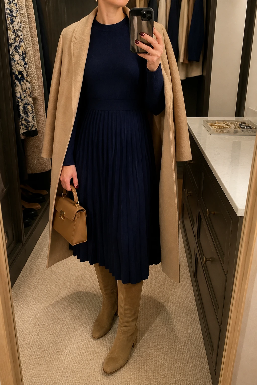 Image Prompt: Mirror selfie of a woman wearing a navy long-sleeve pleated wool-blend midi dress with a modest high neckline and defined waist, tan suede knee-high boots with low block heels, a long beige wool wrap coat worn open, a small structured tan leather handbag, and small pearl stud earrings, holding a phone in front of a mirror, cropped at the NECK so the face does not show, with a relaxed casual selfie pose, in a walk-in closet mirror, warm soft indoor lighting, an open closet with clothes visible and a jewelry tray on the vanity, realistic navy pleated dress fabric, natural tan suede texture and soft coat folds, latest iPhone photo quality, no additional clothing or accessories.